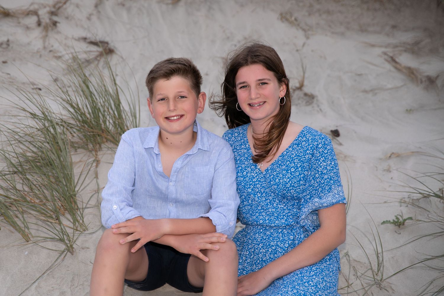 Two people in coordinating blue and white outfits sit together on a sandy beach during a photo session.