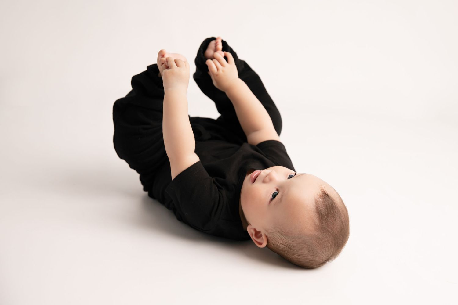 Baby wearing black onesie lies on back against white background while playing with feet.
