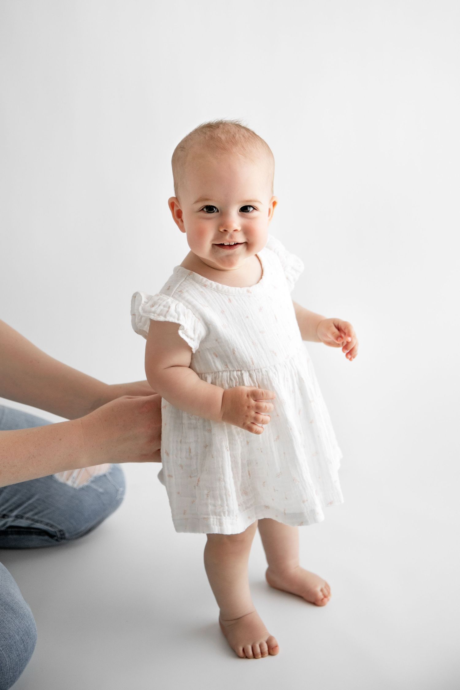 Baby in white eyelet dress learning to walk with support on minimalist white background.