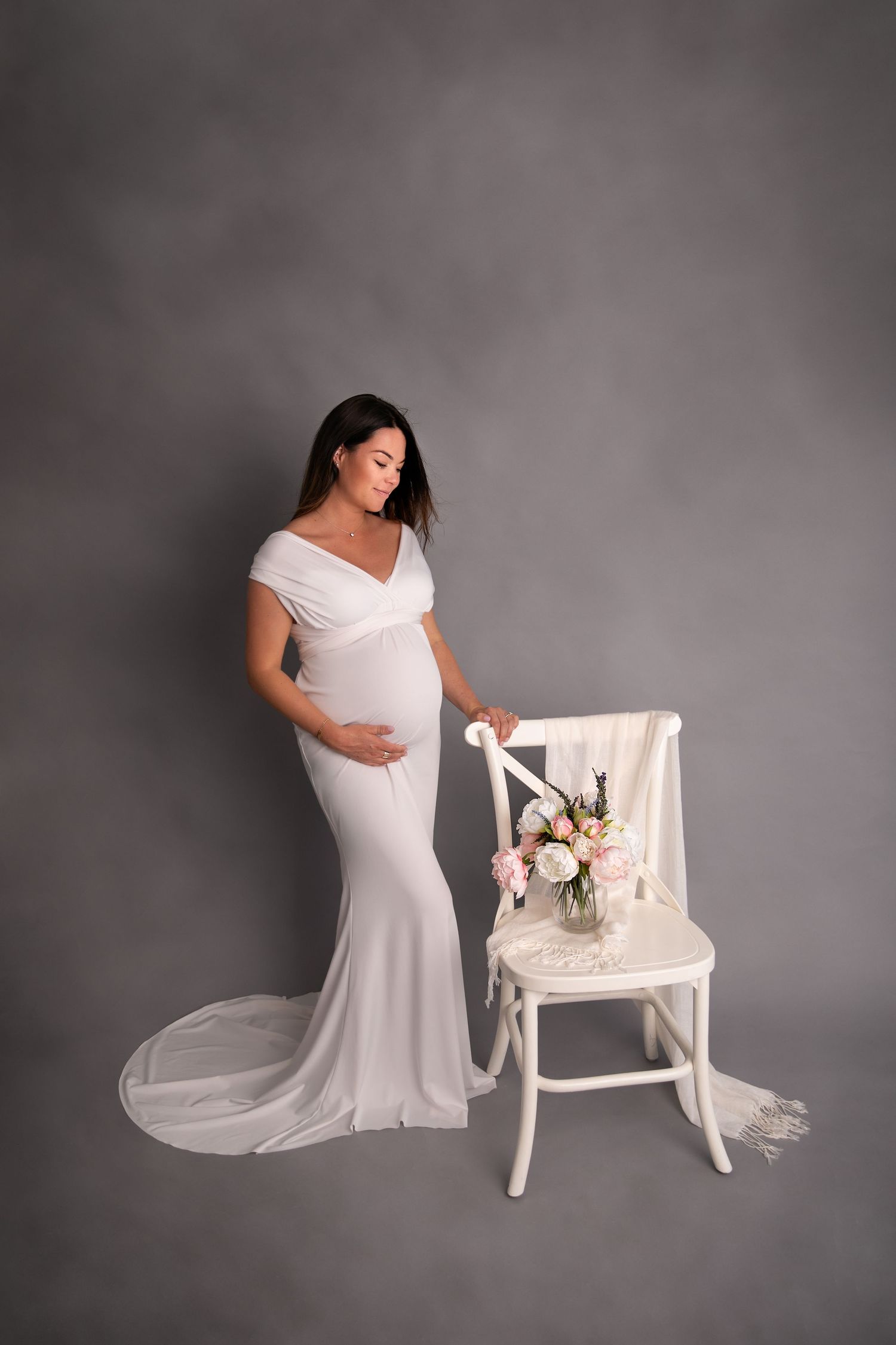 A pregnant woman in a flowing white gown stands beside a white chair adorned with floral decorations against a gray backdrop.