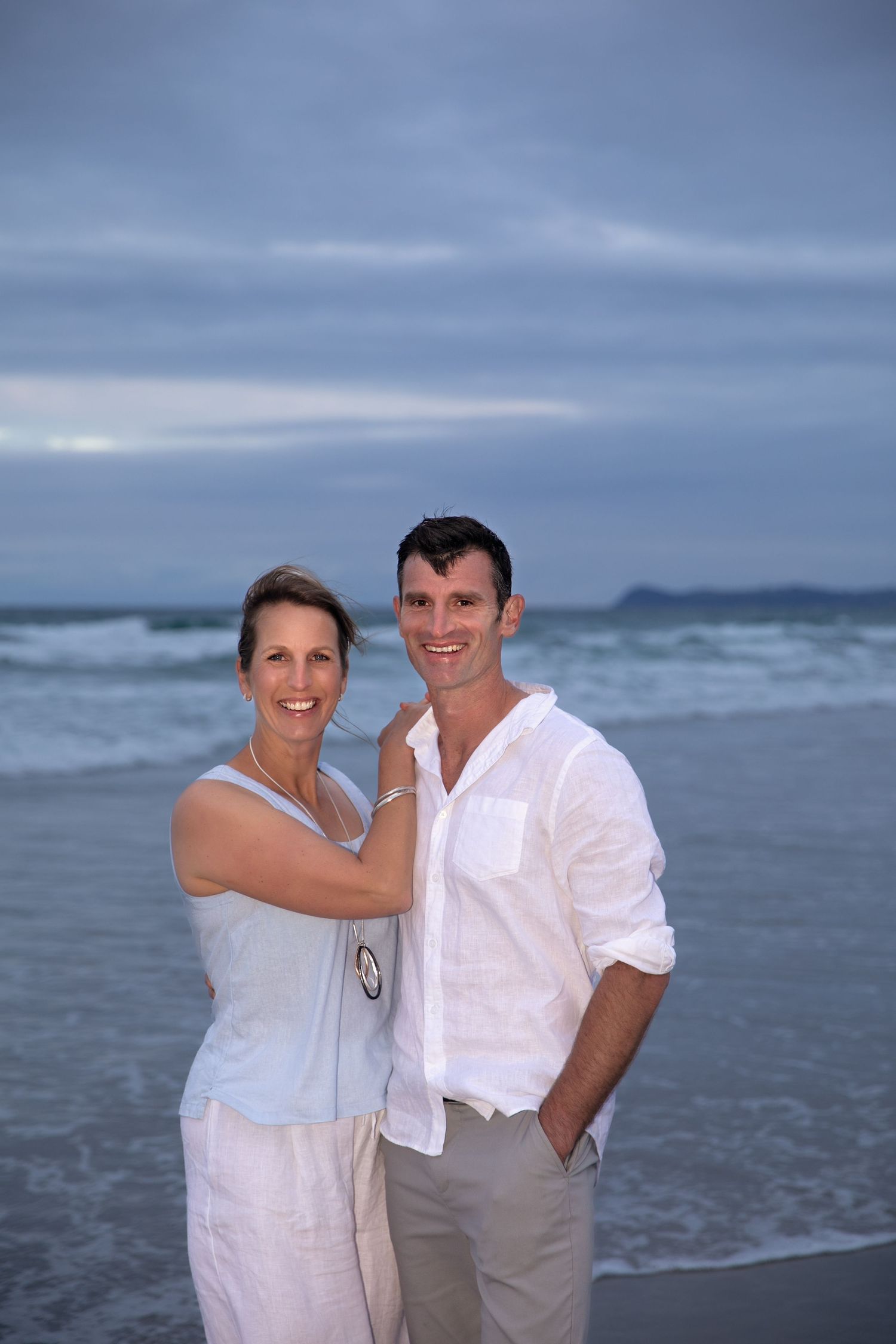 A couple dressed in white stands together on a beach at dusk with ocean waves and cloudy sky in the background.