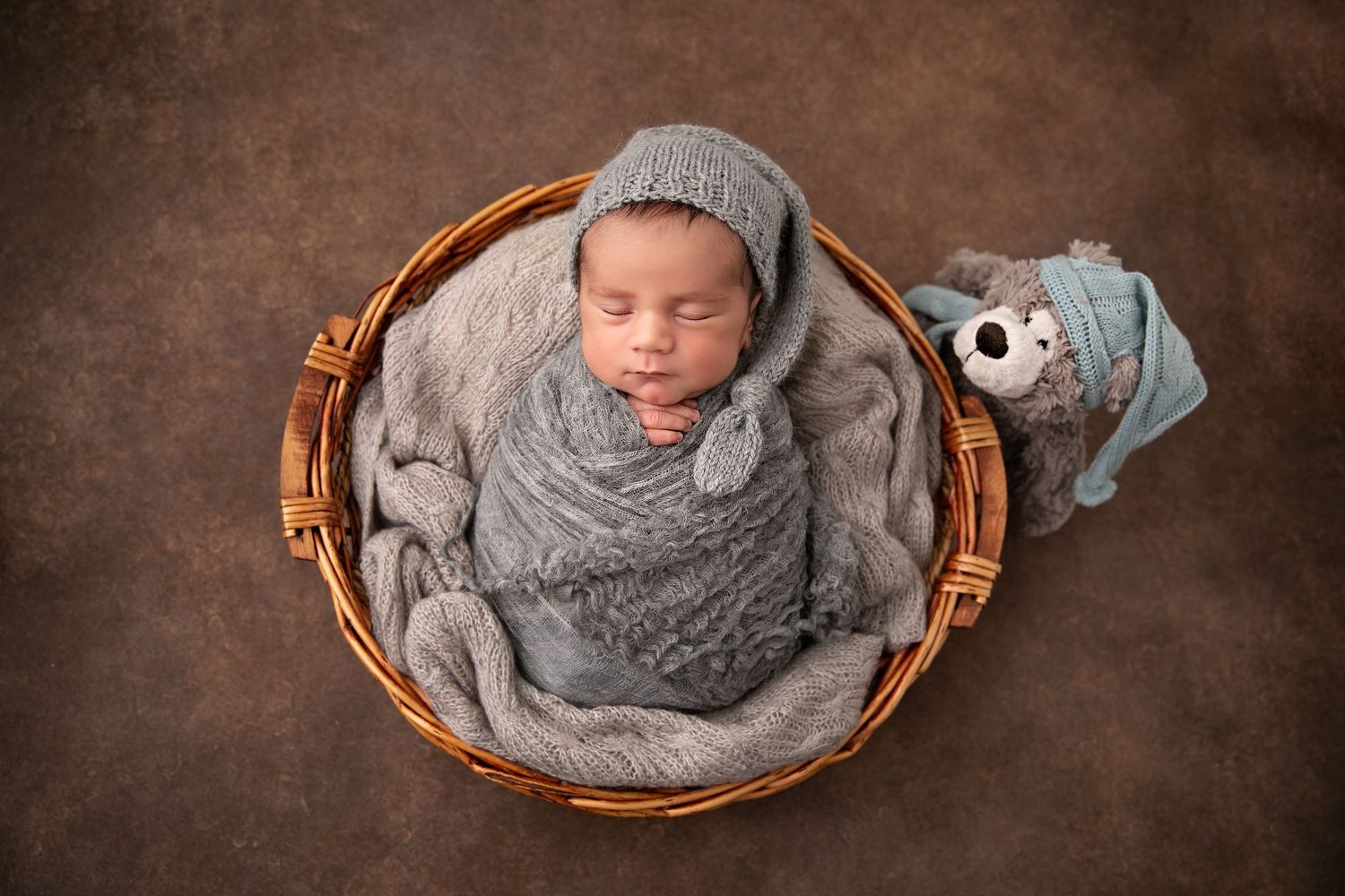 A sleeping newborn baby swaddled in soft gray fabric lies peacefully in a wooden basket against a brown backdrop.
