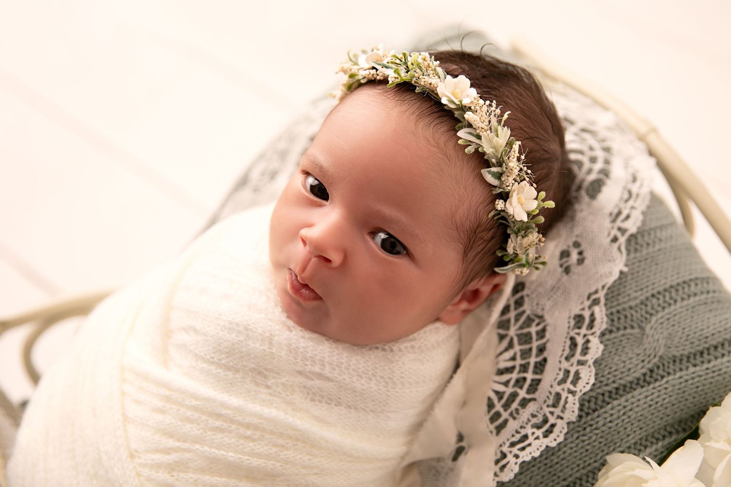 A newborn baby wearing a delicate white floral crown and wrapped in soft gray fabric lies peacefully on a light background.