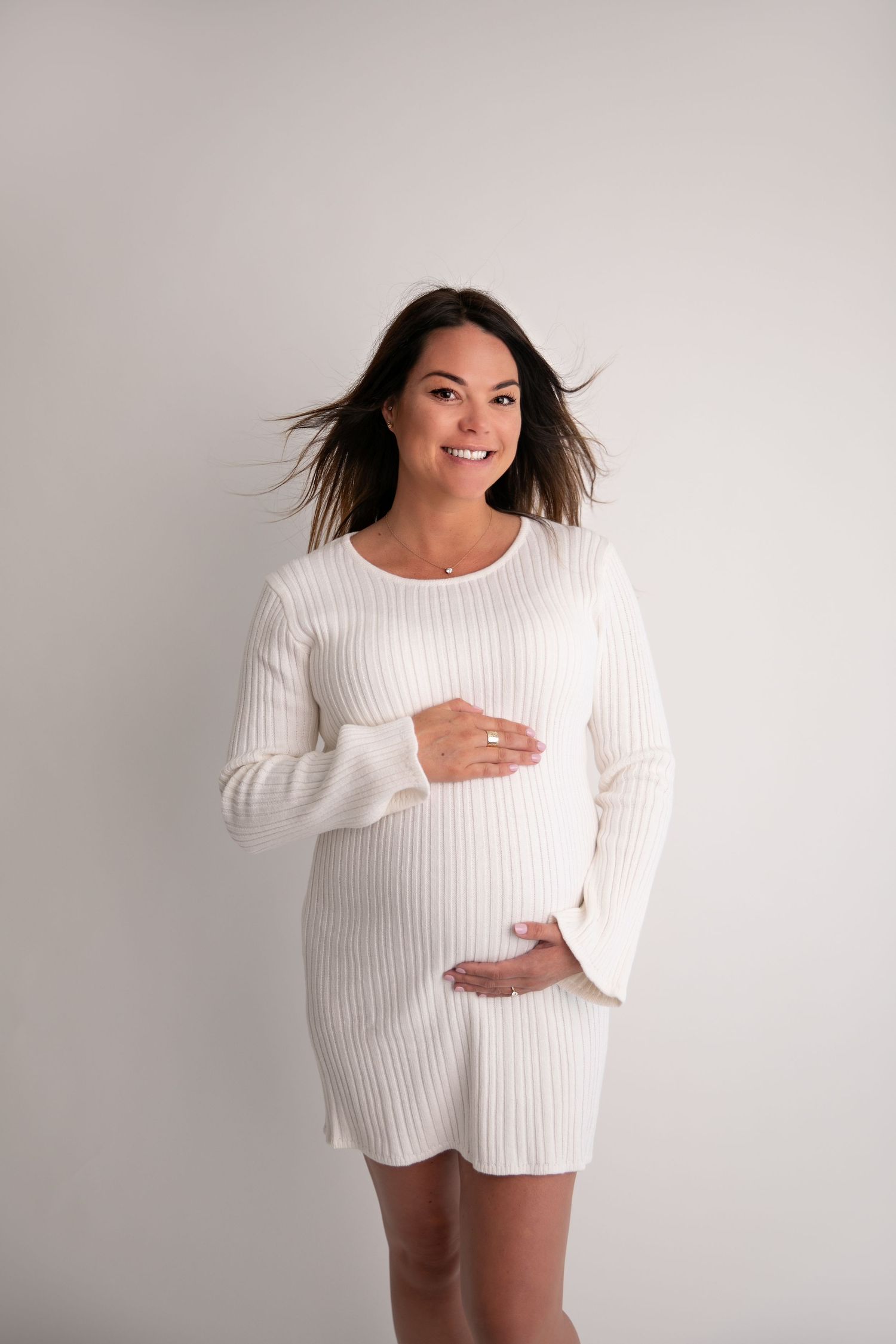 Expectant woman in white ribbed sweater dress poses against light background with wind-blown hair.