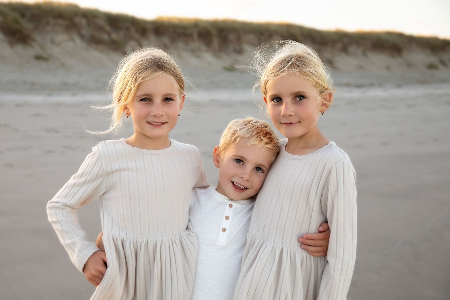 Two children wearing white outfits pose together on a sandy beach at sunset.