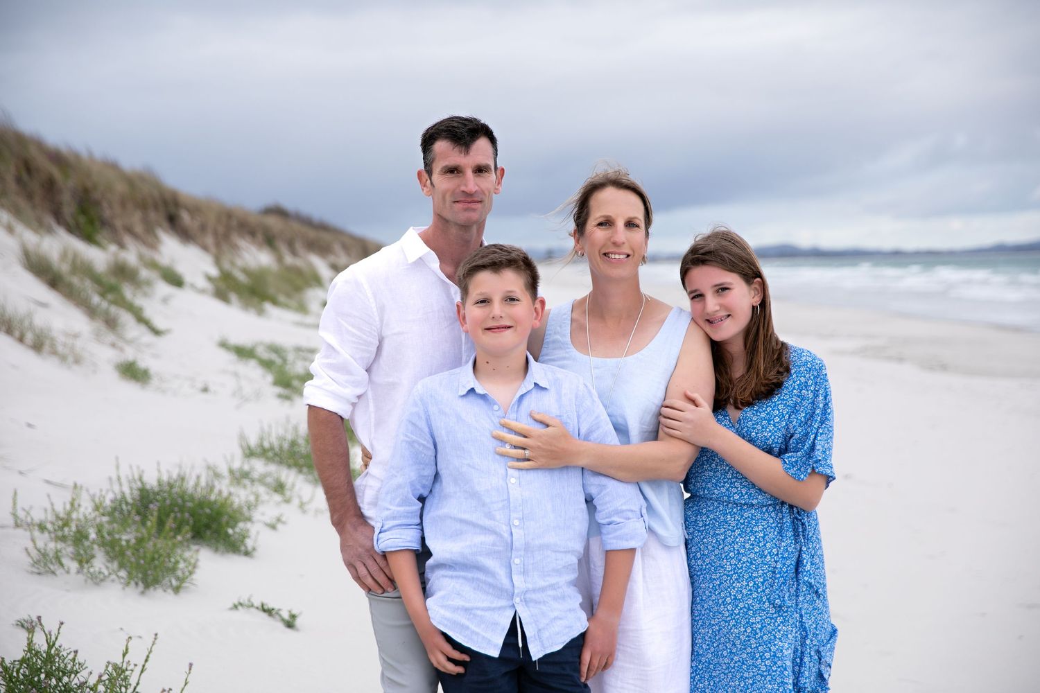 Family wearing coordinated blue and white outfits poses together on sandy beach with ocean waves in background.