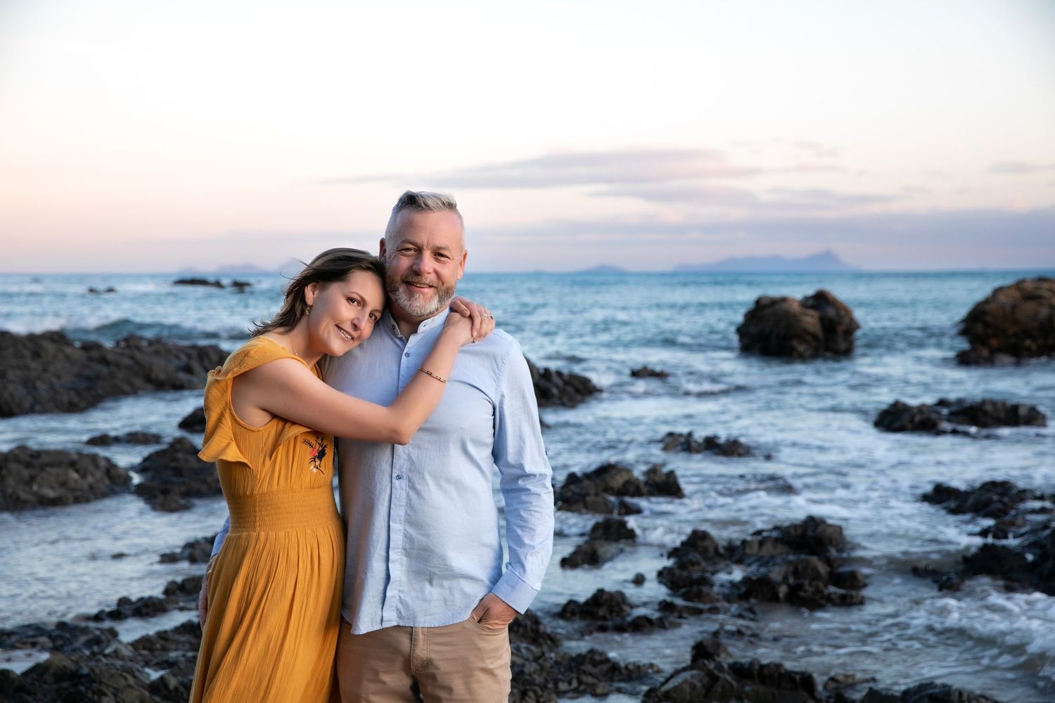Couple embracing on rocky beach at sunset with ocean waves and coastal rocks in background.