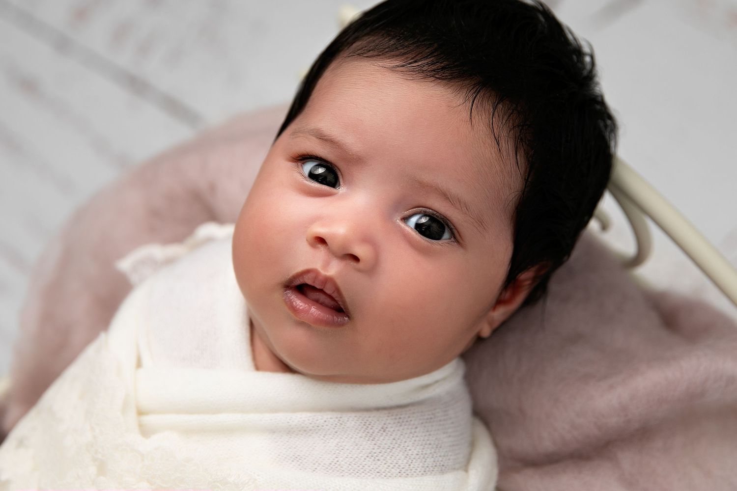 A baby wearing a white onesie lies on a light gray blanket while looking up at the camera.