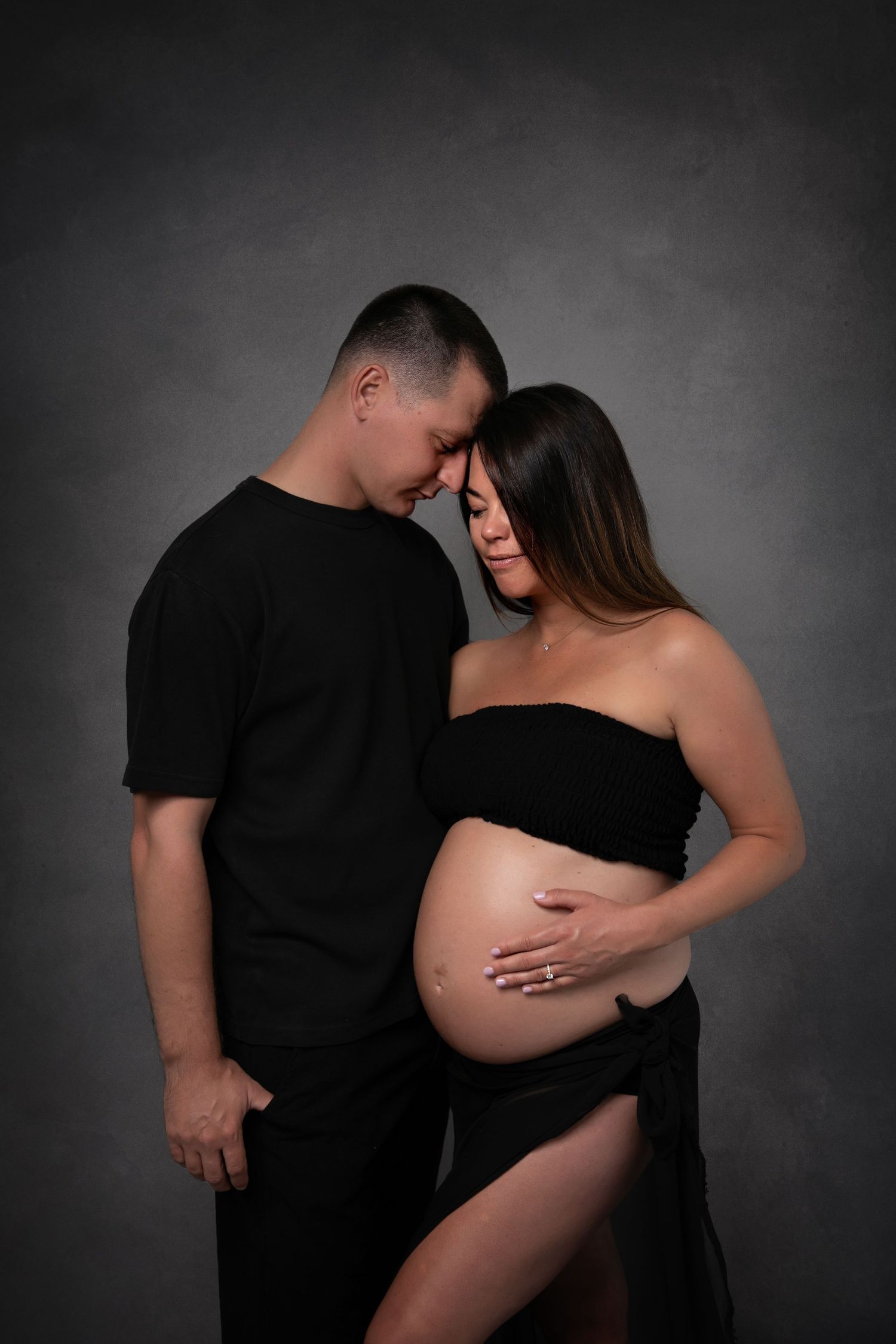 Artistic black and white maternity photo series showing intimate moments between expectant parents in dark studio setting.