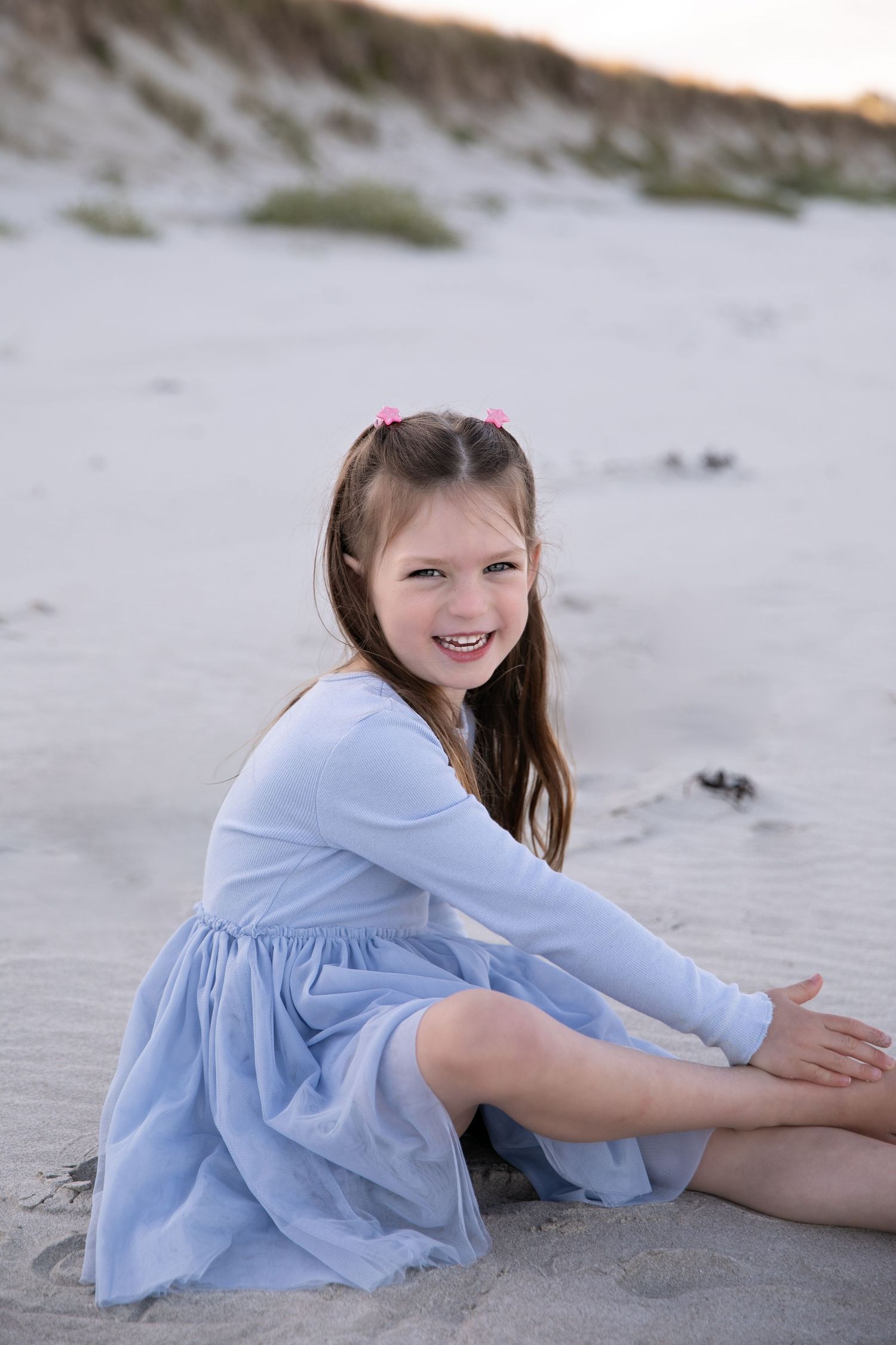 Child in a light blue dress sits on a sandy beach at sunset with a joyful expression.