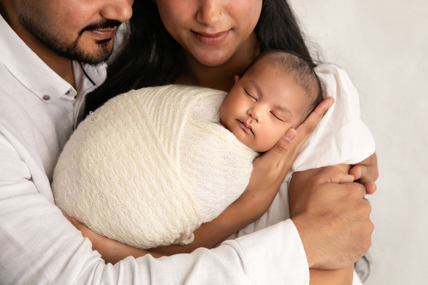 A sleeping newborn baby swaddled in white is held lovingly by parents in a tender family portrait.