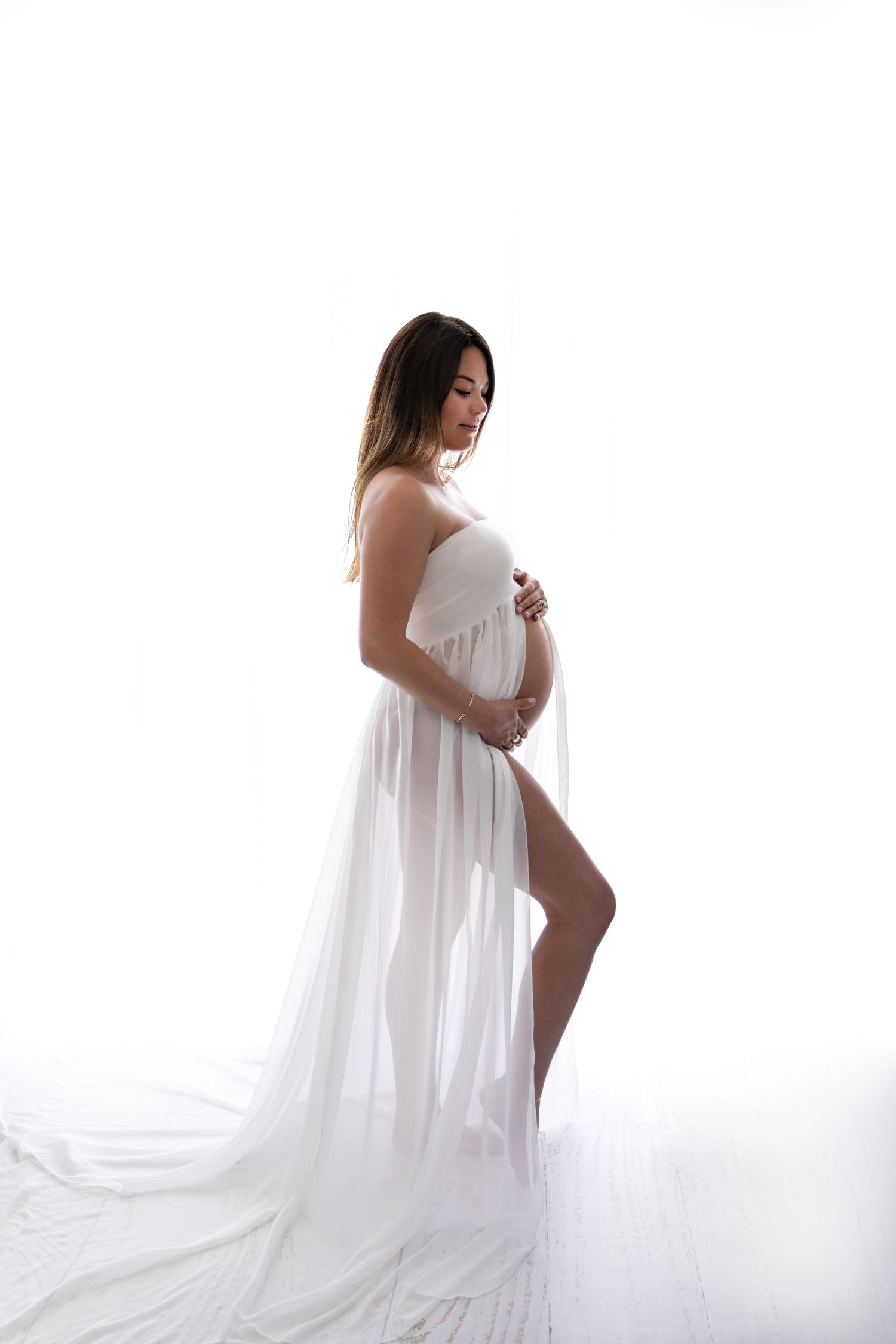 Artistic maternity photoshoot featuring a woman in flowing white dress against bright white background.