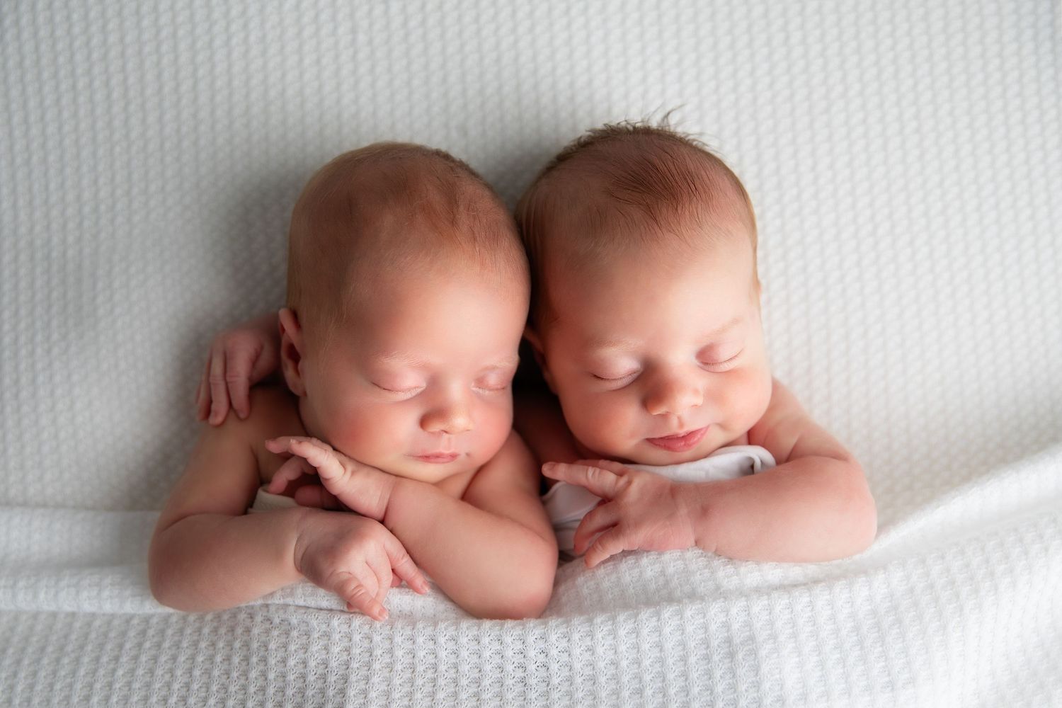 Two sleeping newborn babies lying side by side on soft white bedding in a peaceful pose.