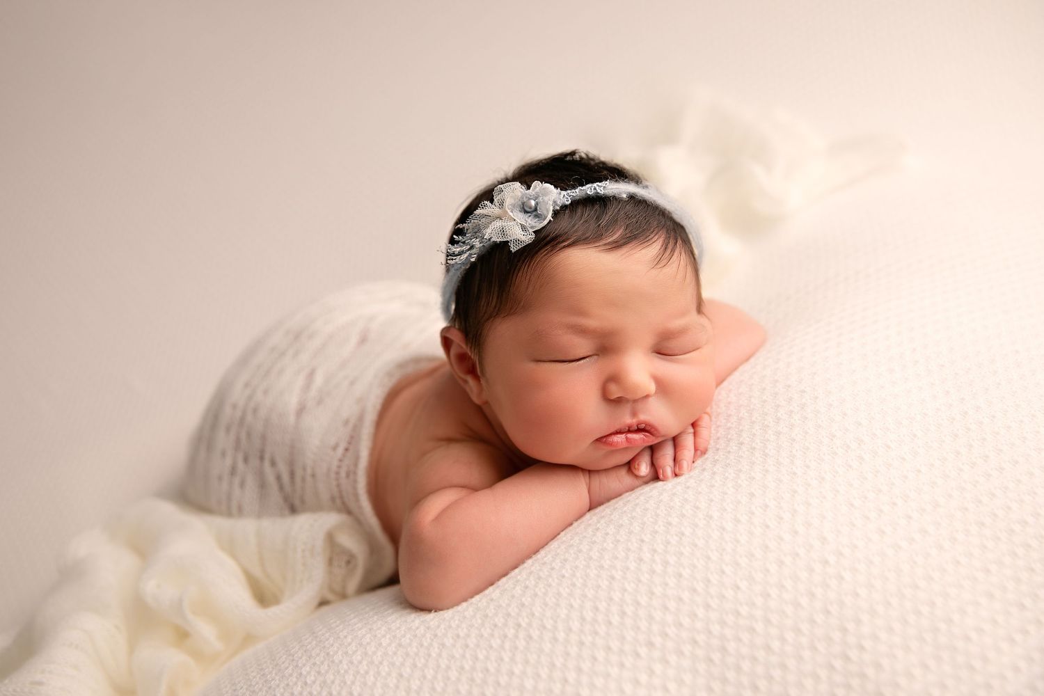 Sleeping newborn baby in delicate white wraps and headbands on soft cream backdrop.