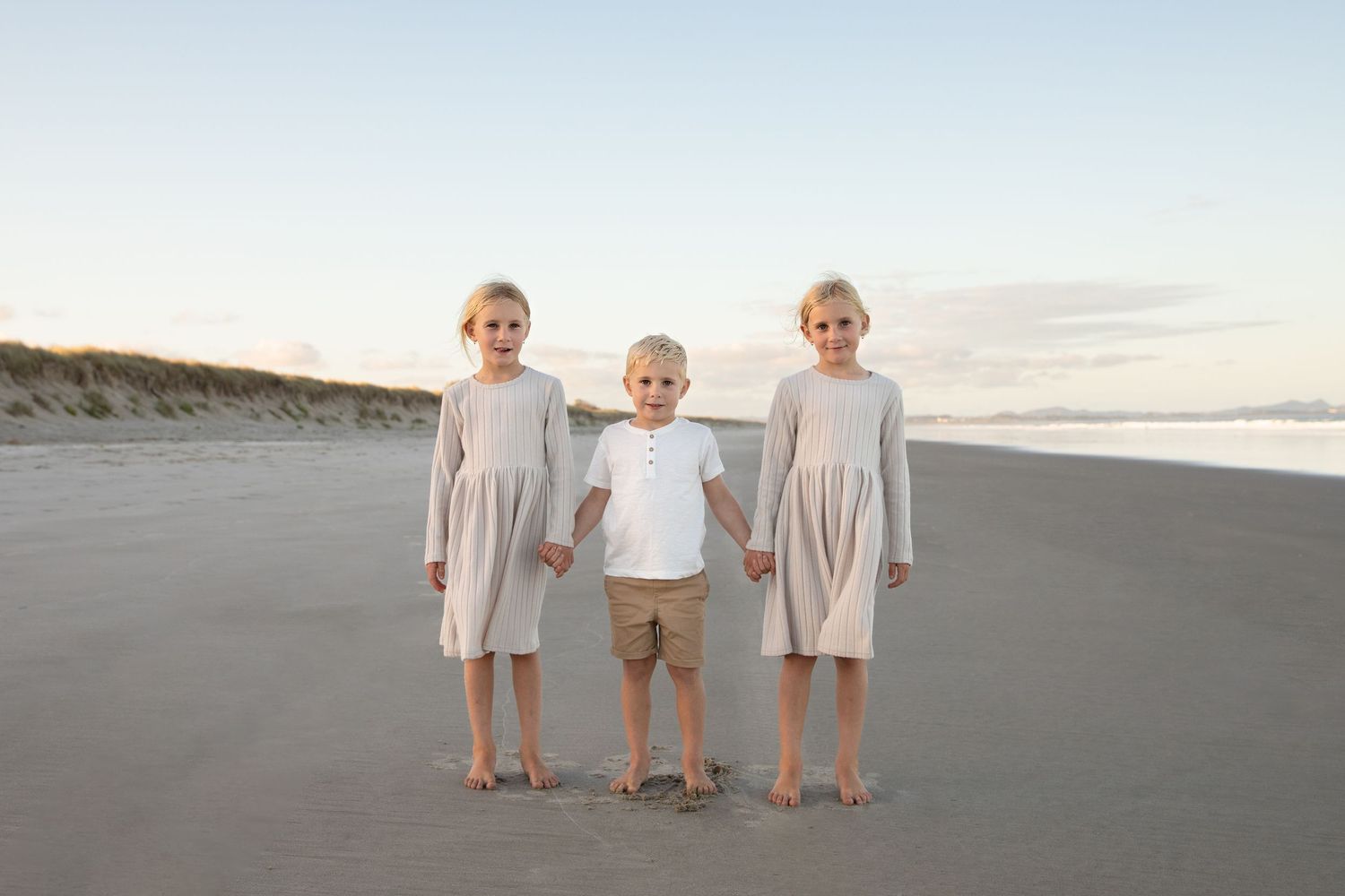 Three young children walk hand in hand along a sandy beach at sunset wearing coordinated neutral-colored outfits.