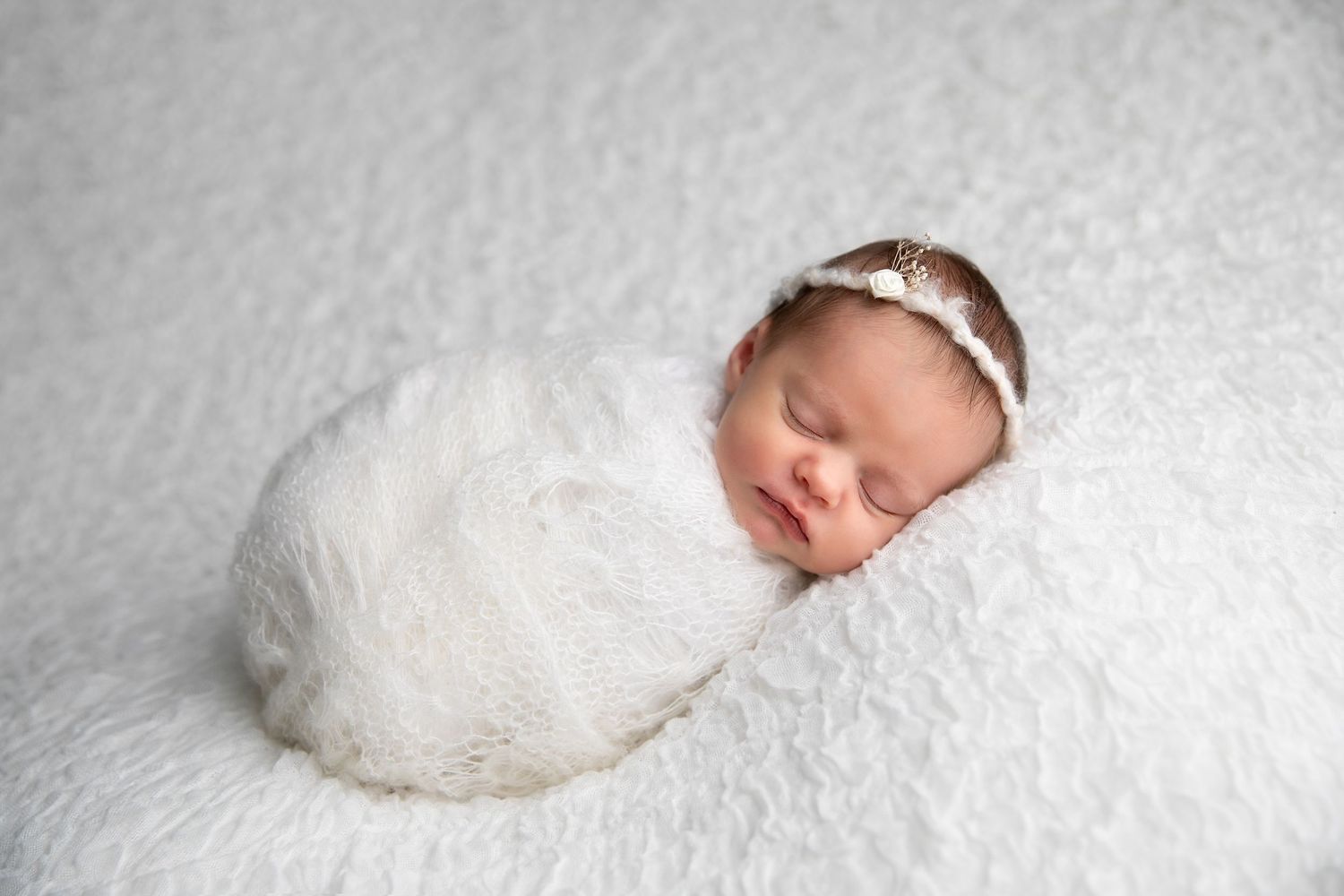 A sleeping newborn baby wrapped in soft white fabric against a light gray textured background.