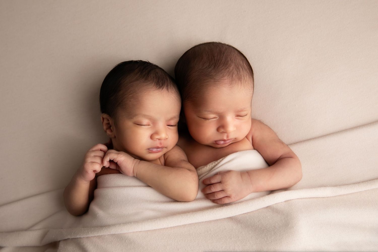 Two sleeping newborn babies snuggled close together on soft white fabric in a tender newborn photography portrait.