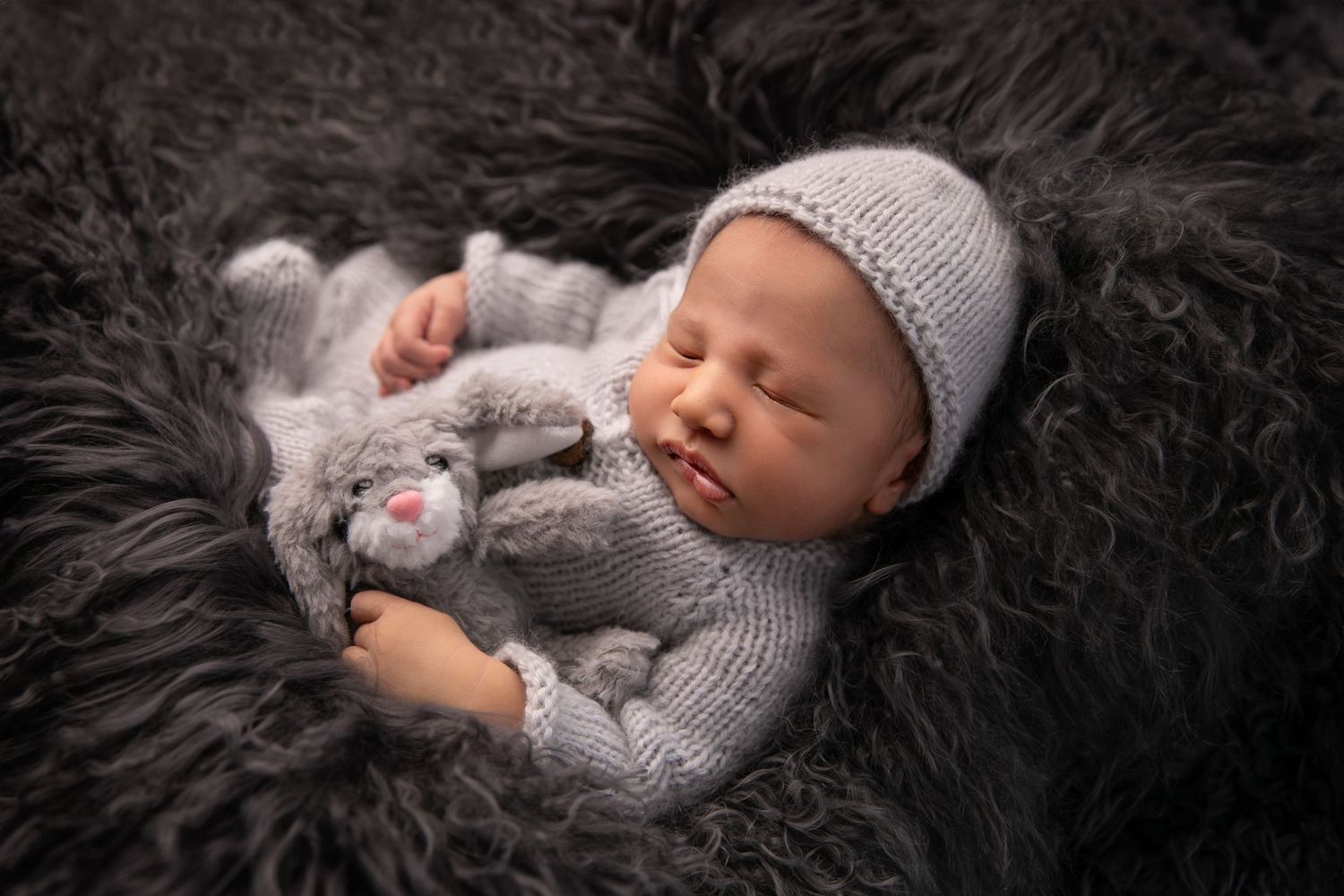 A sleeping baby wearing a gray knit outfit lies on a plush black fur blanket with a small stuffed animal.