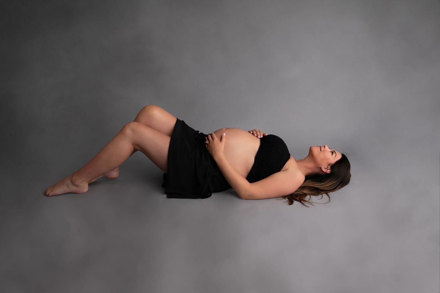 A person in a black dress lies on a gray backdrop in an artistic studio pose.