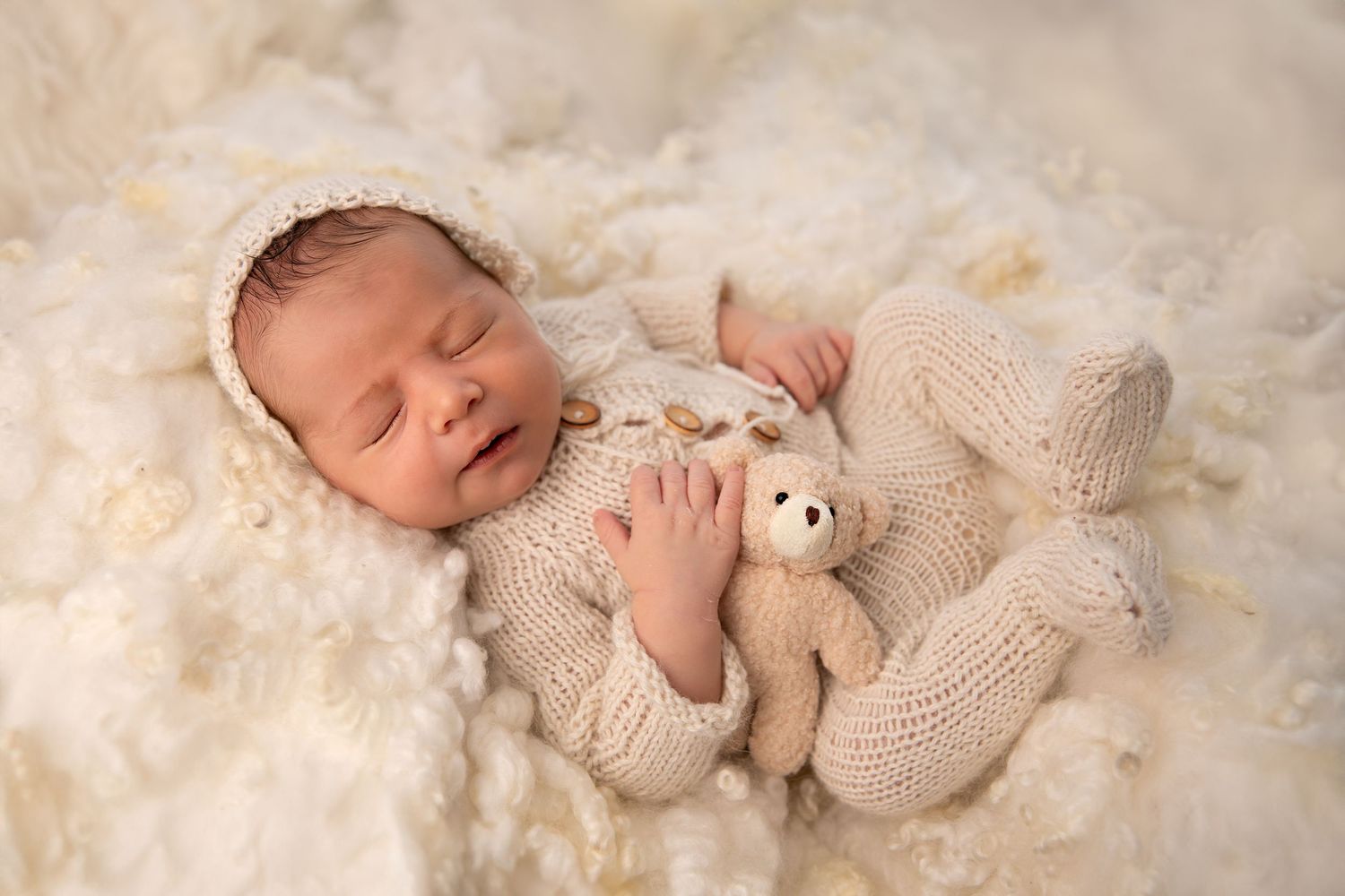 Newborn baby sleeping peacefully on soft cream lace blanket with small teddy bear toy.