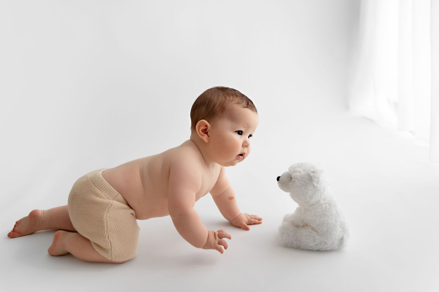 A baby in a diaper crawls curiously toward a white teddy bear on a light background.
