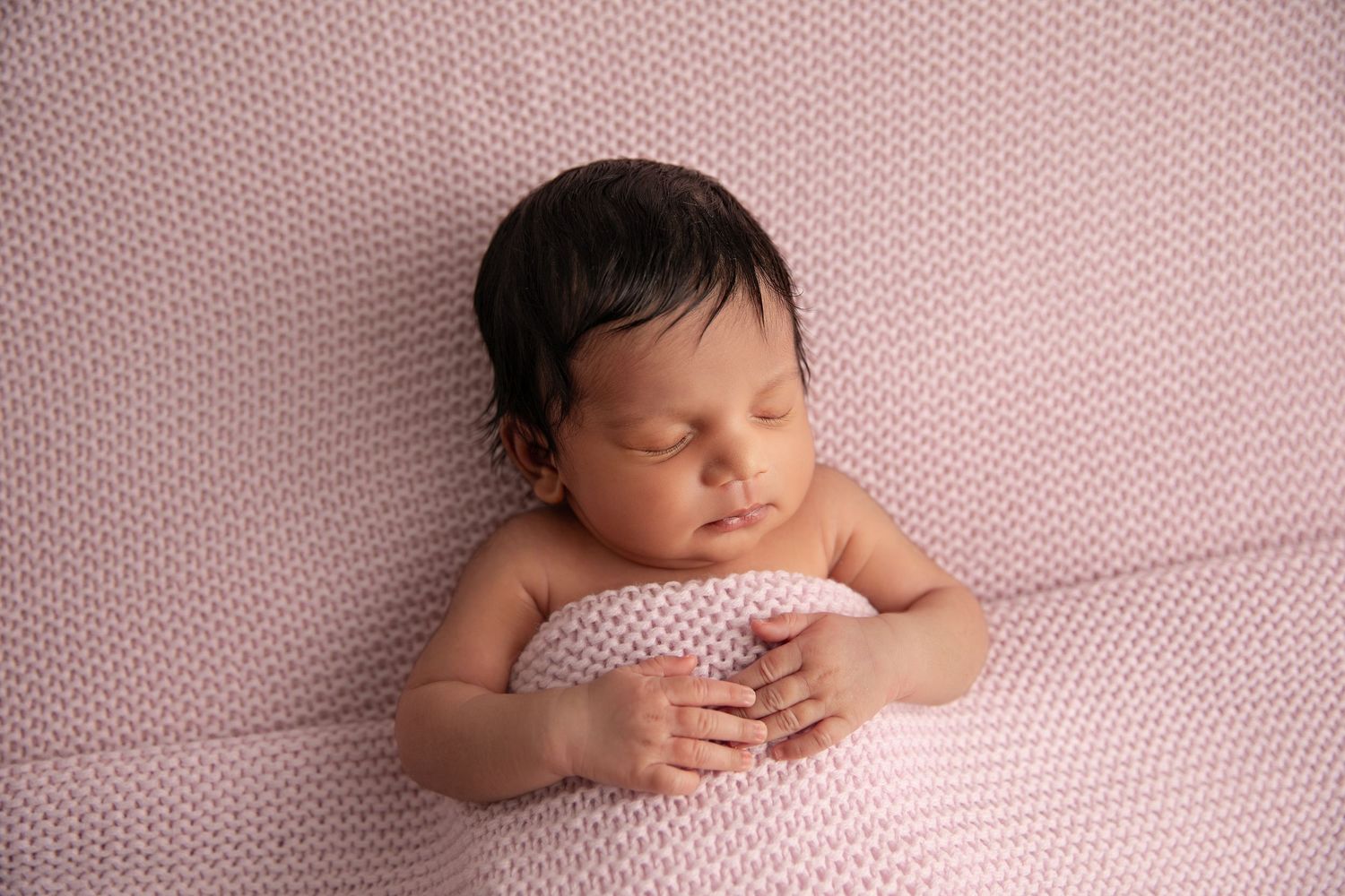 Sleeping newborn baby wrapped in pink textured fabric lying on matching pink blanket.