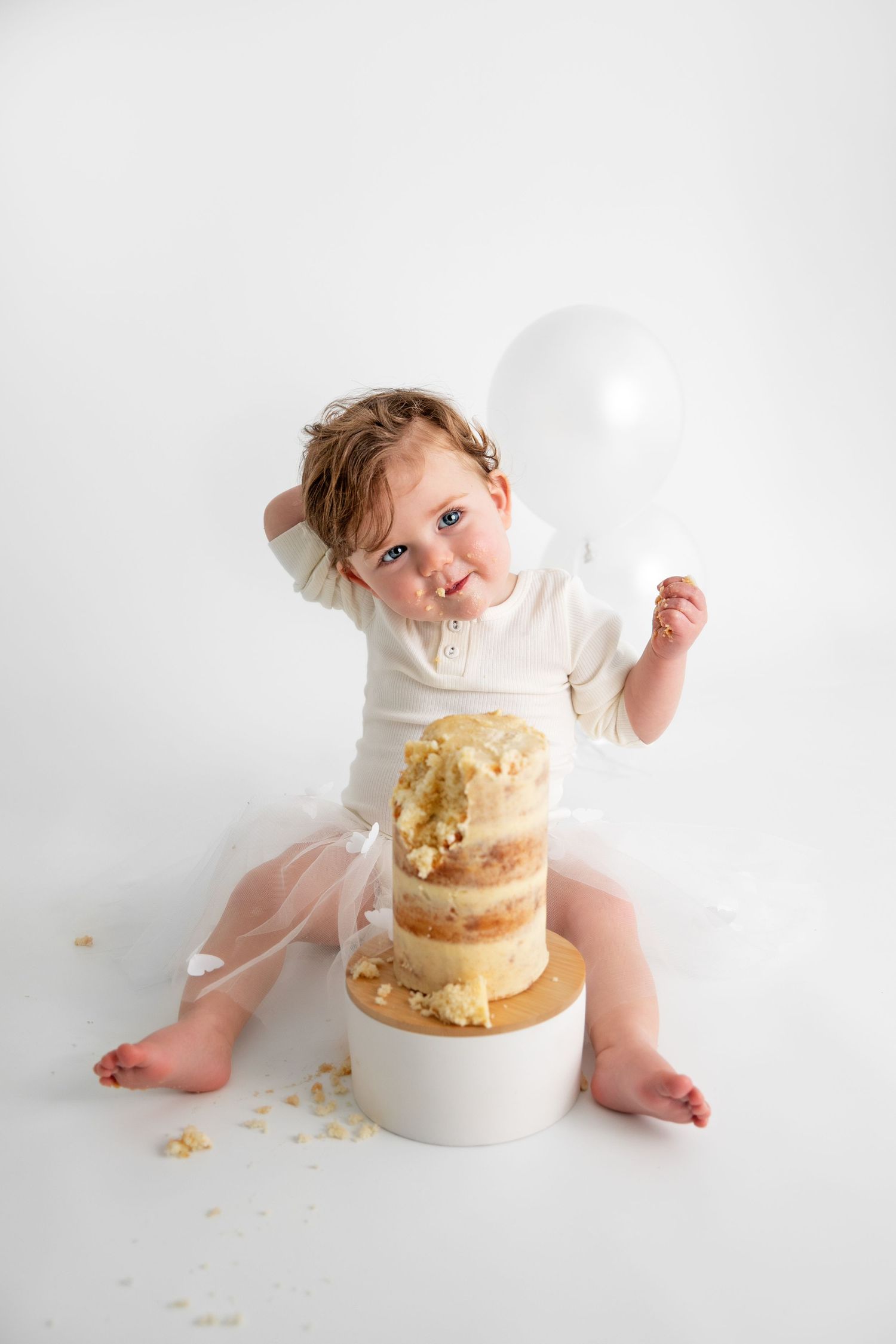 Baby in white outfit sits beside elegant gold and white tiered cake during cake smash photo session.