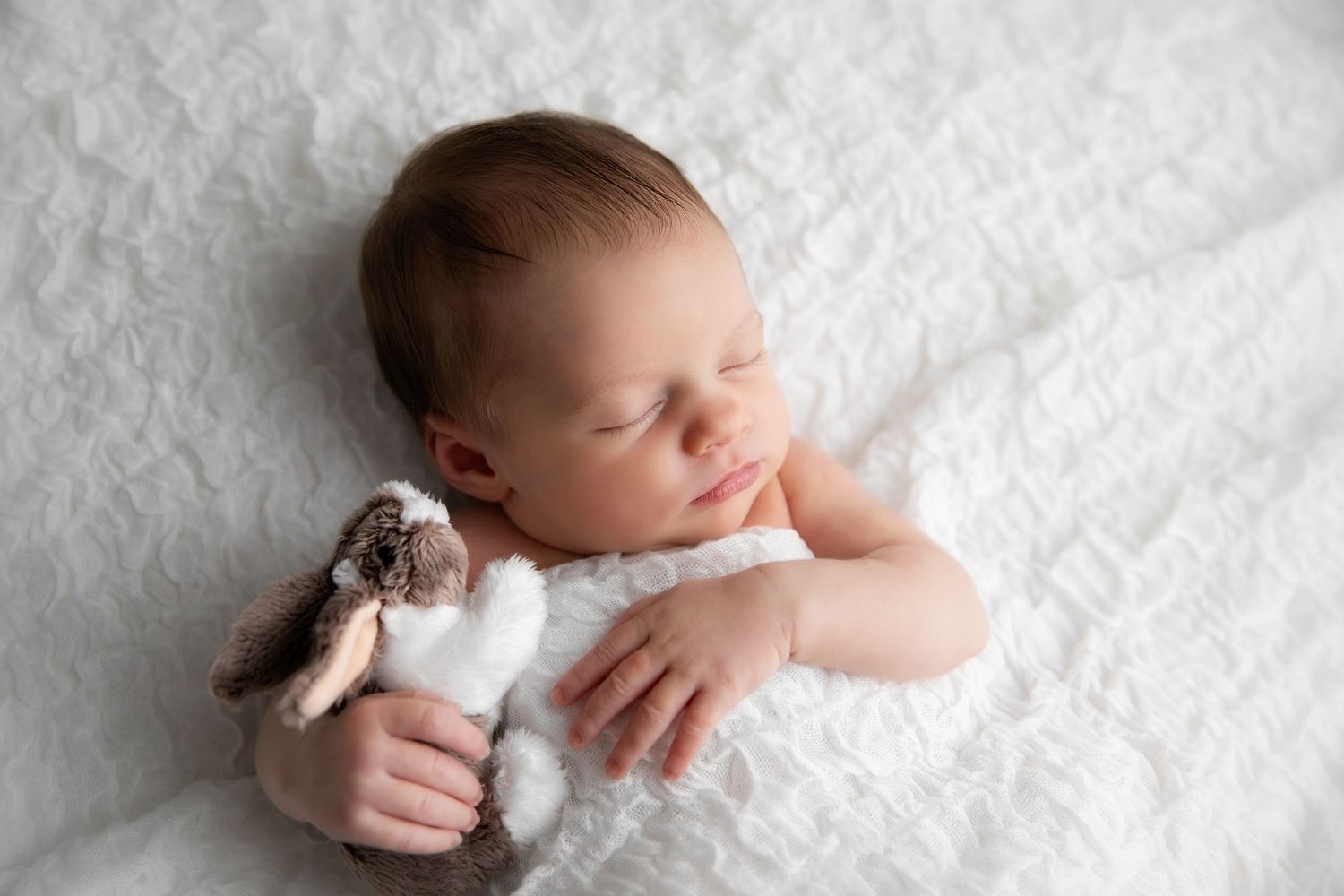 Sleeping newborn in white swaddle cuddles with small plush bunny toy on soft white blanket.