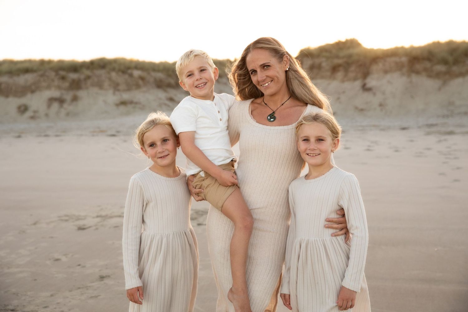 A family in coordinating beige outfits poses together on a sandy beach at sunset.