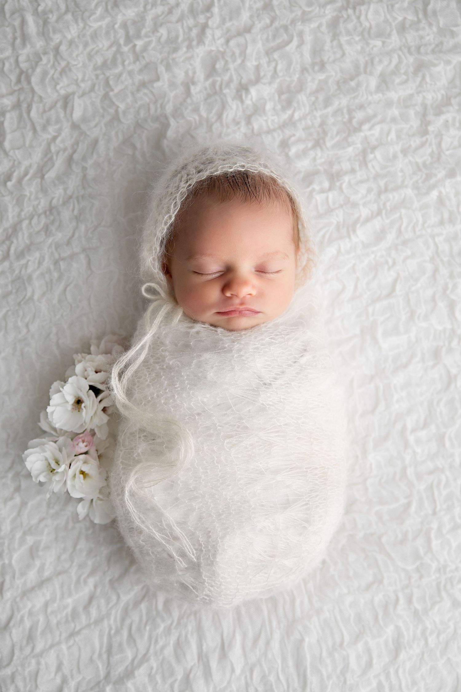 Sleeping newborn swaddled in soft white fabric with delicate floral accents on textured white background.