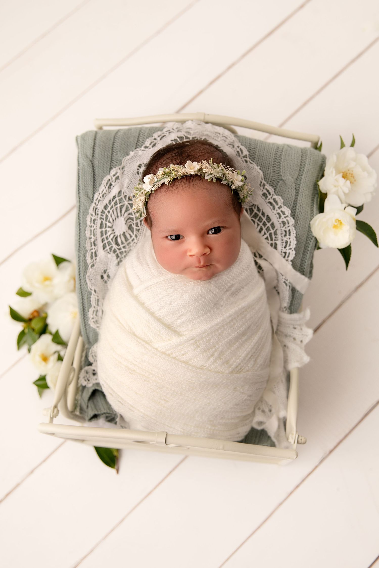A newborn baby swaddled in white fabric with delicate lace details lies peacefully beside white flowers on a wooden backdrop.