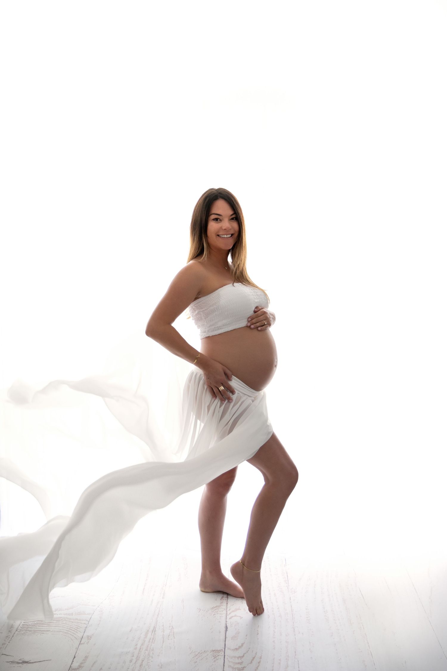 Ethereal maternity photo of an expectant mother in flowing white fabric against a bright minimalist backdrop.