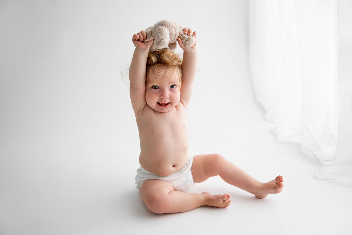 Happy baby wearing a white diaper sits and plays on a bright white backdrop.