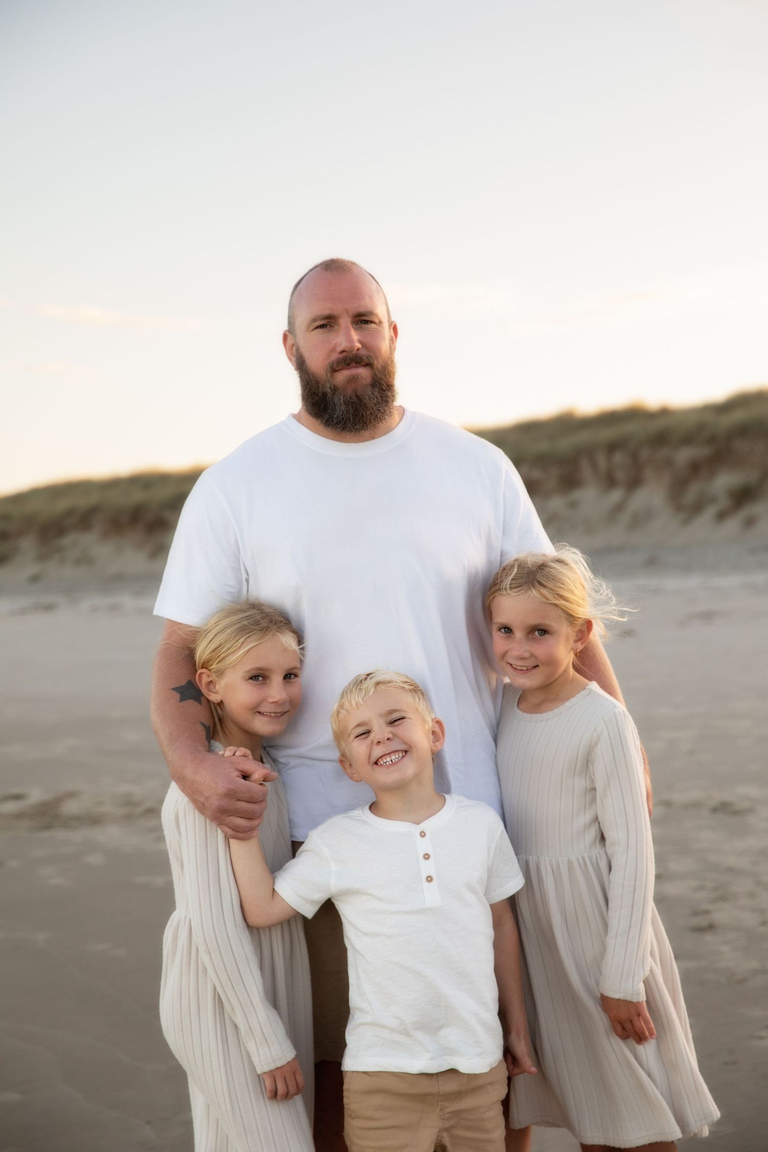 A family stands together on a sandy beach at sunset wearing white and neutral colored clothing.