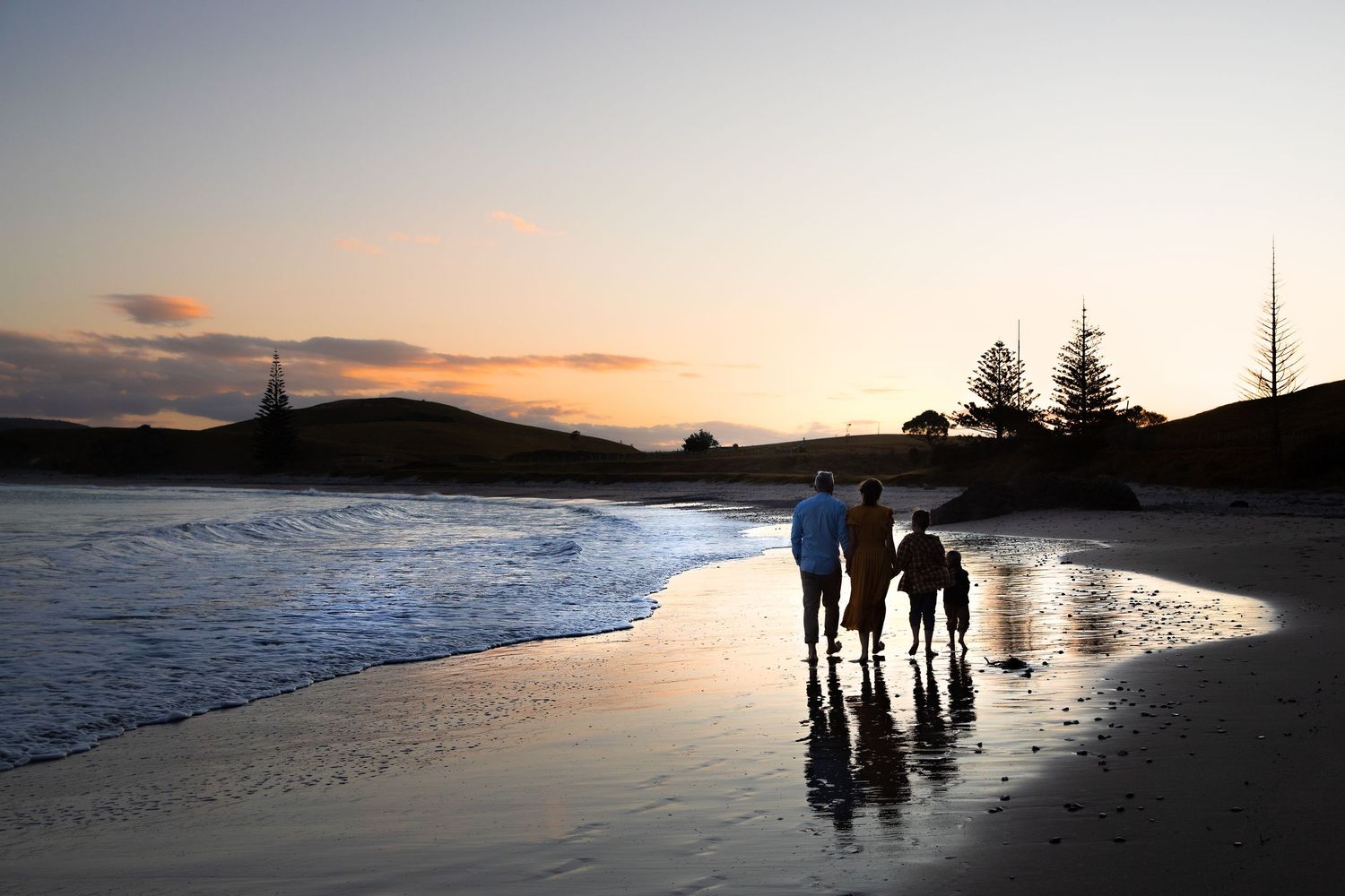 Silhouettes of beachgoers stroll along a scenic coastline at sunset with waves rolling onto the shore.