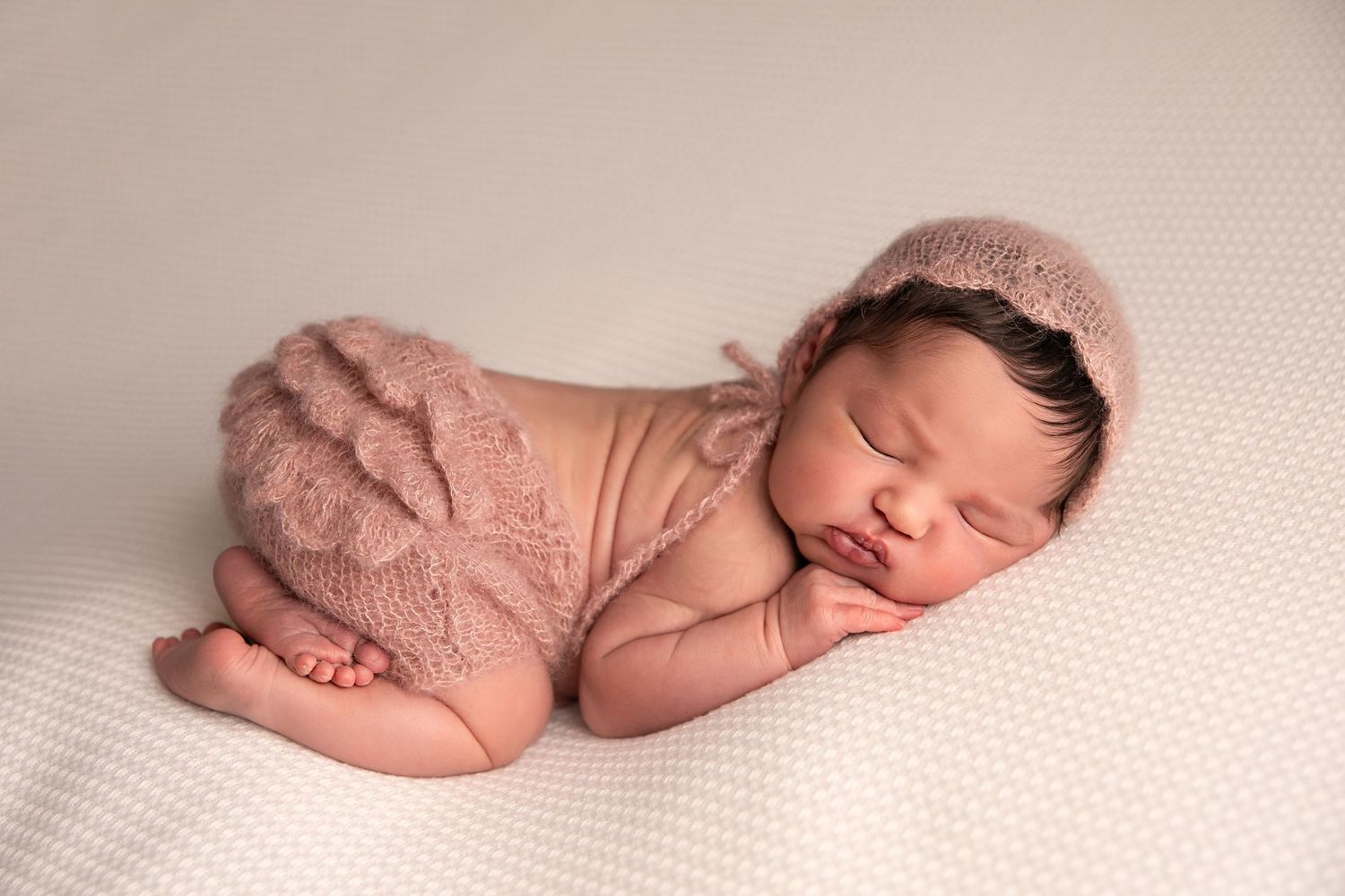 Sleeping newborn baby wearing pink lace romper rests peacefully on white textured backdrop during professional photoshoot.