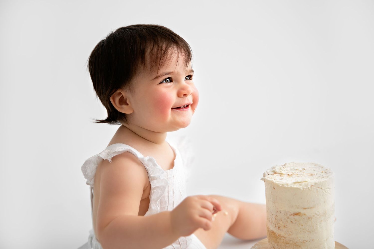 Baby in white dress reaches for birthday cake with white frosting on white background.