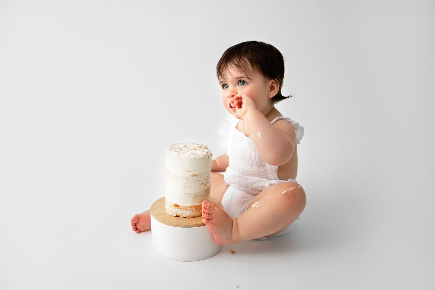 Baby in white dress plays with small cake during photoshoot.