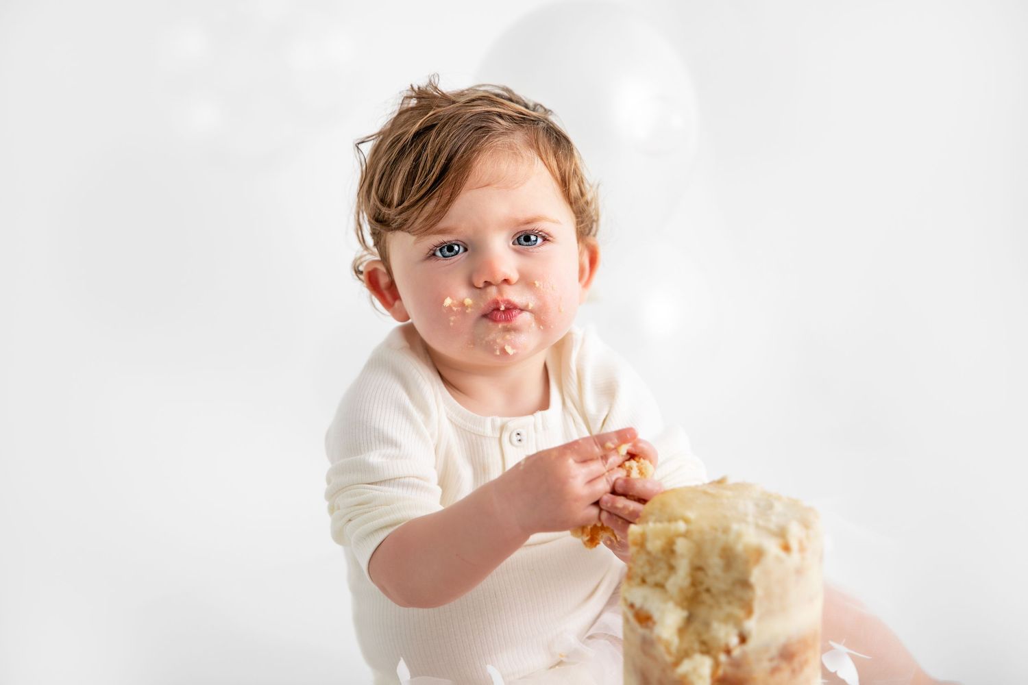 A child in a white dress holds a cream-colored teddy bear against a light background.