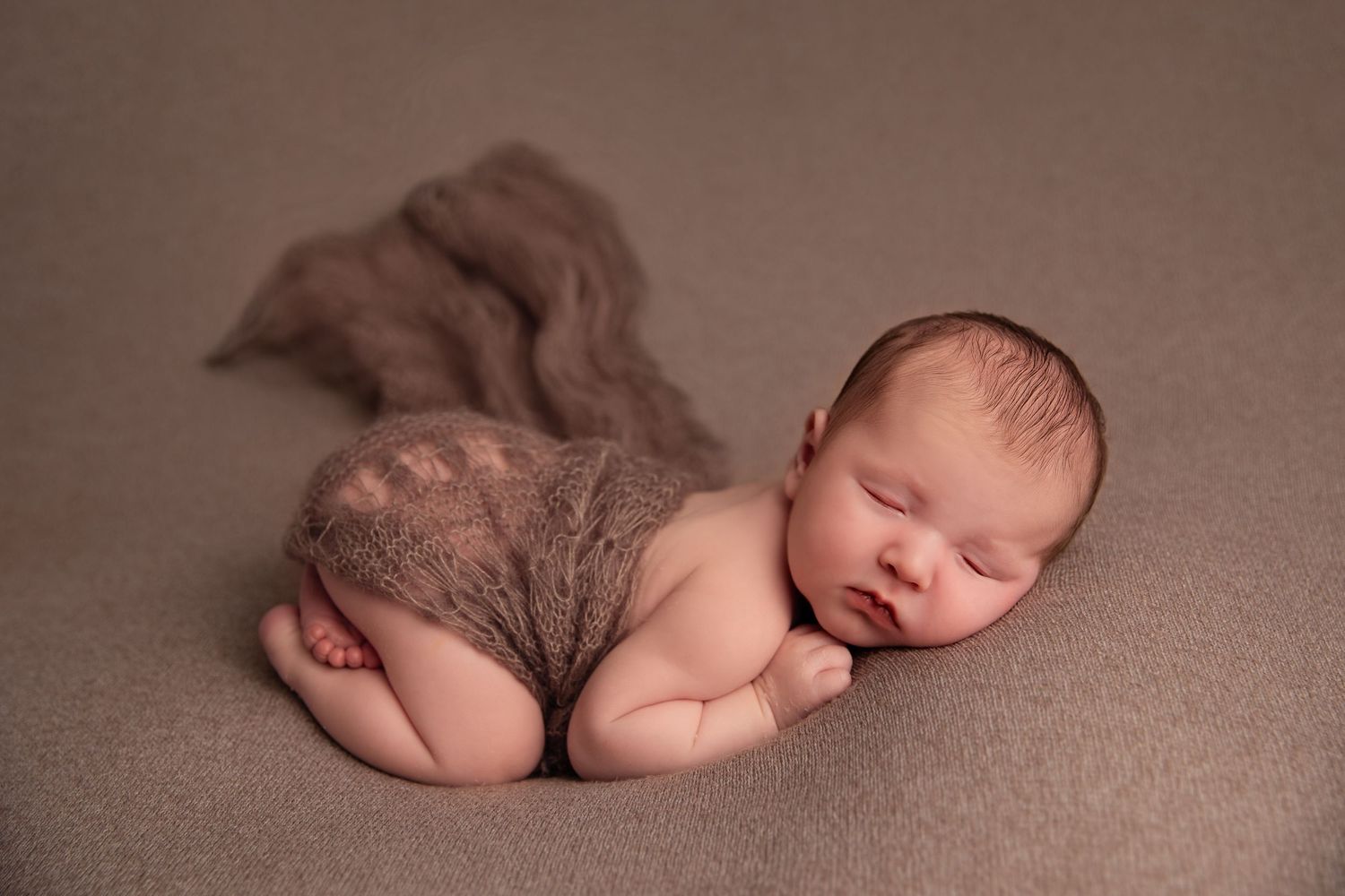 Sleeping newborn baby wearing gray lace diaper cover lying on soft brown backdrop during professional photo session.