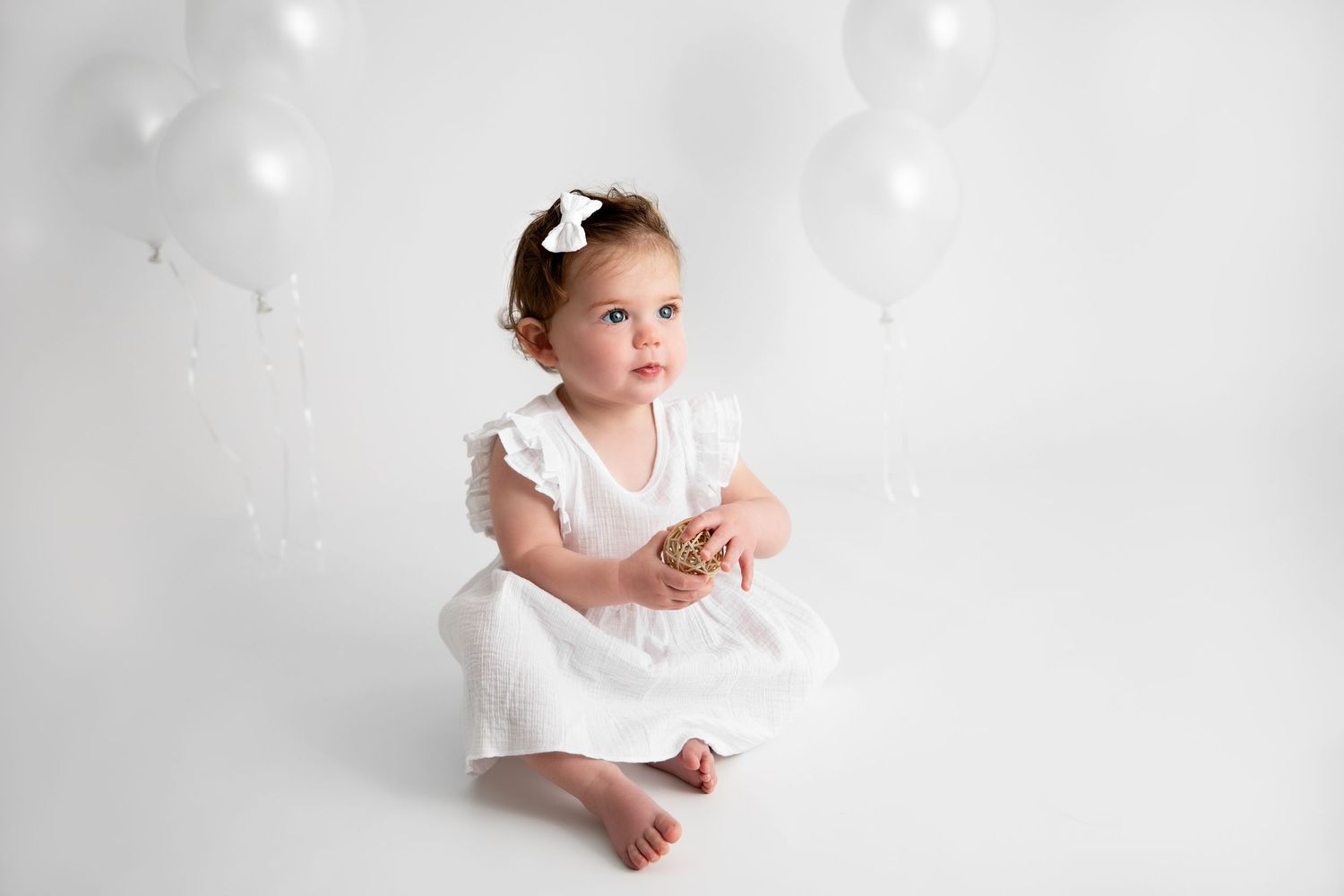 A child in a white dress sits gracefully against a light background in an artistic portrait setting.