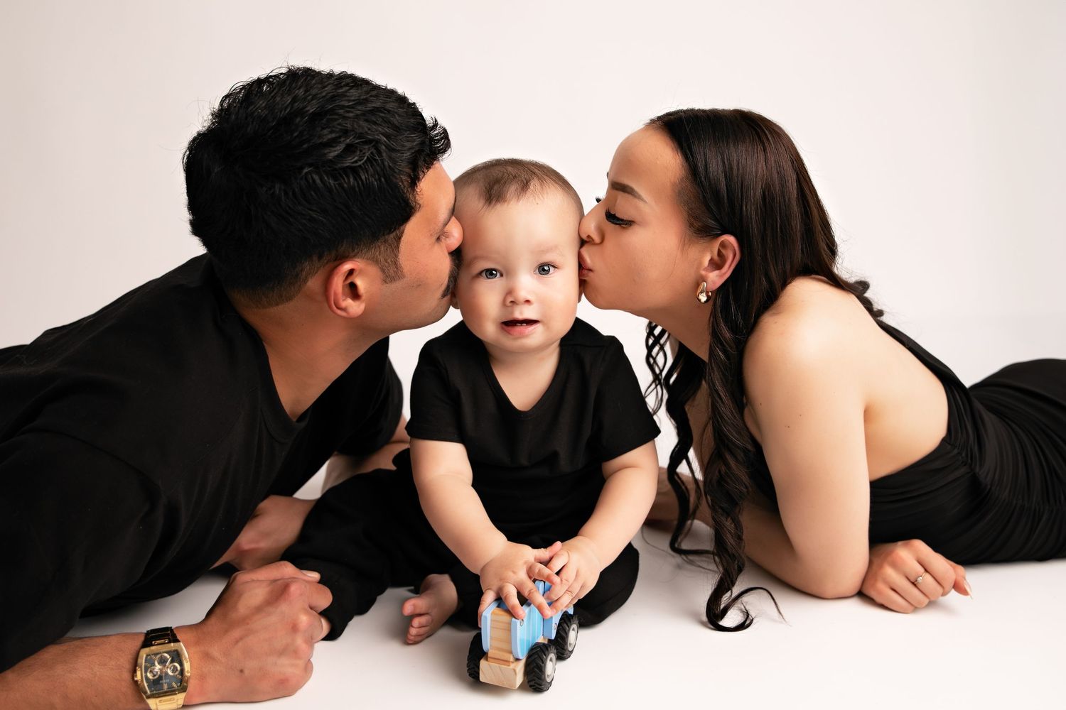 A young child in black clothing sits between two adults on a white background while holding a toy car.