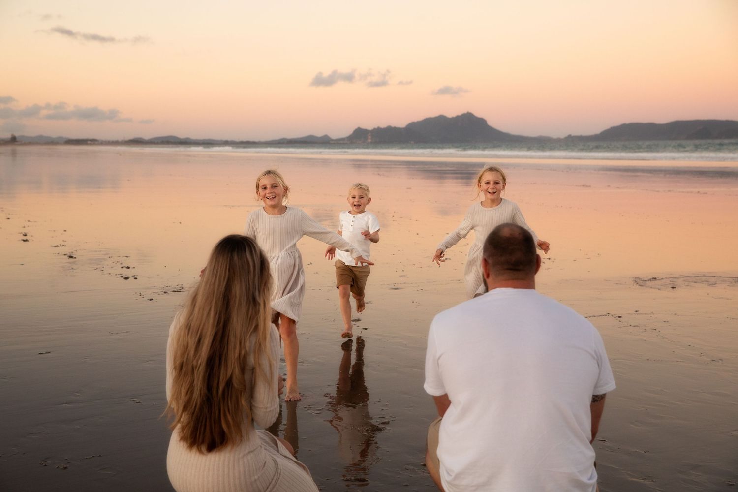 A family enjoys a peaceful sunset moment together on a reflective beach with mountains in the distance.
