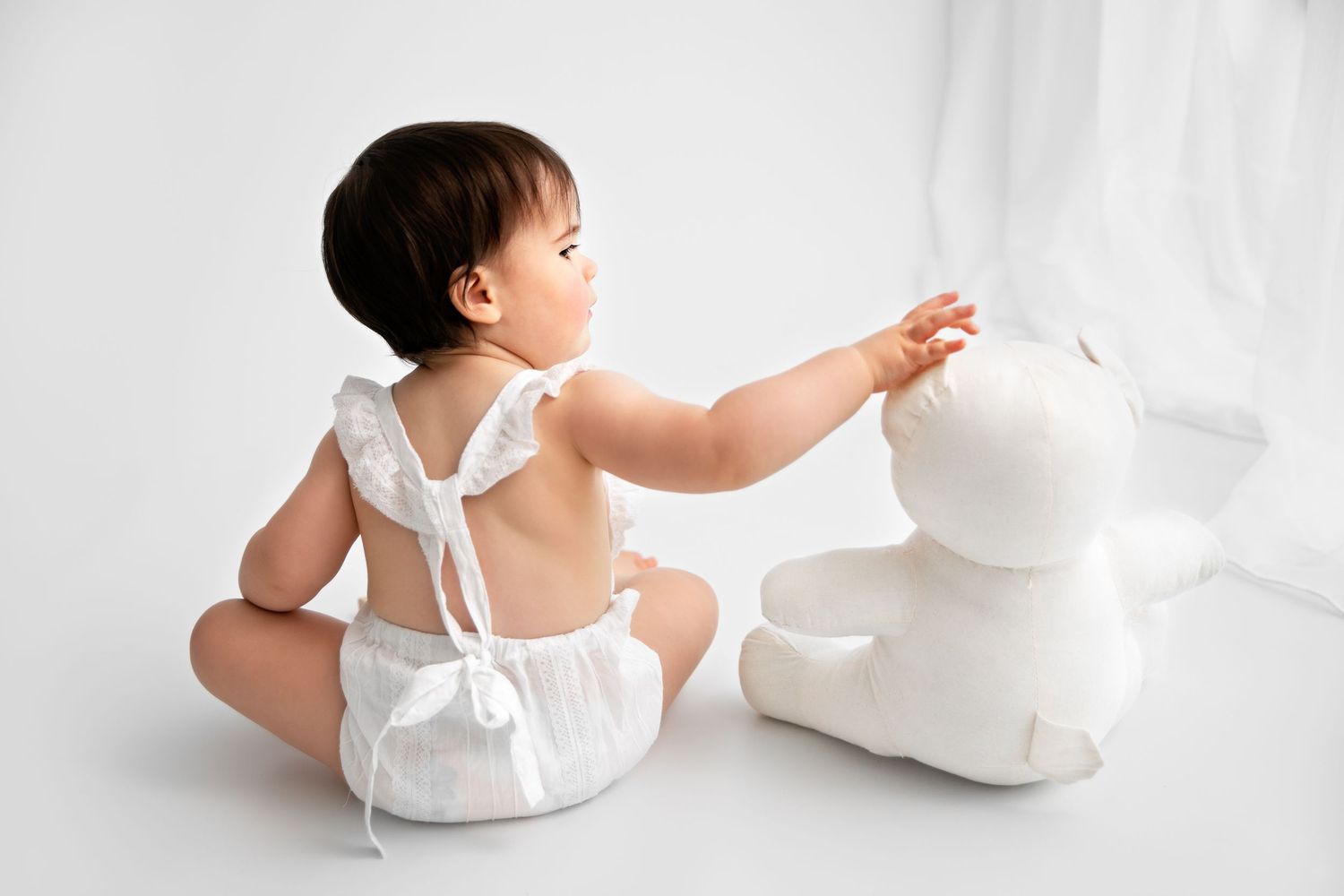 Baby in white dress reaches for cake during studio photoshoot.