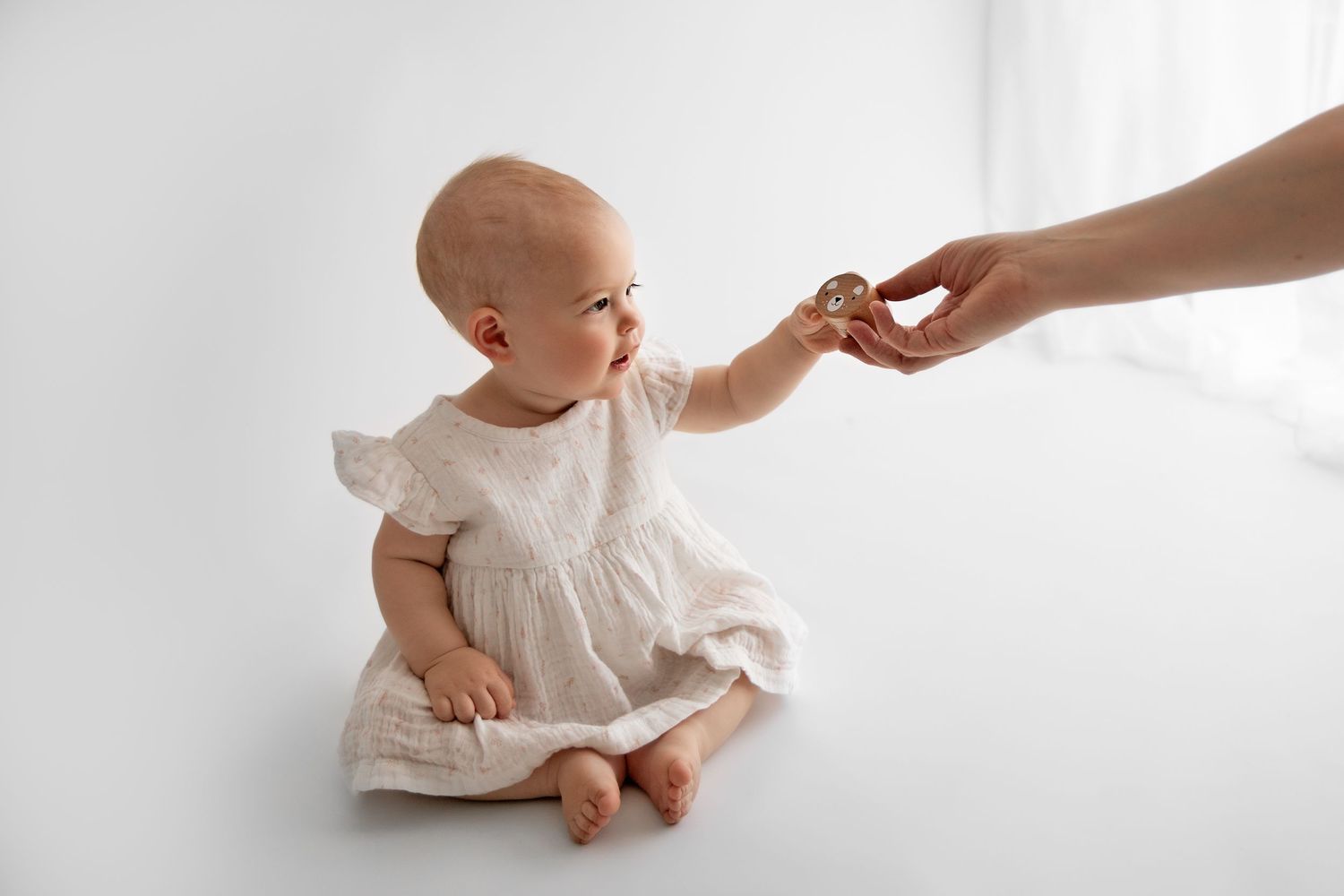 A gentle hand reaches out to comfort a small baby wearing white clothing while sitting on a white surface.