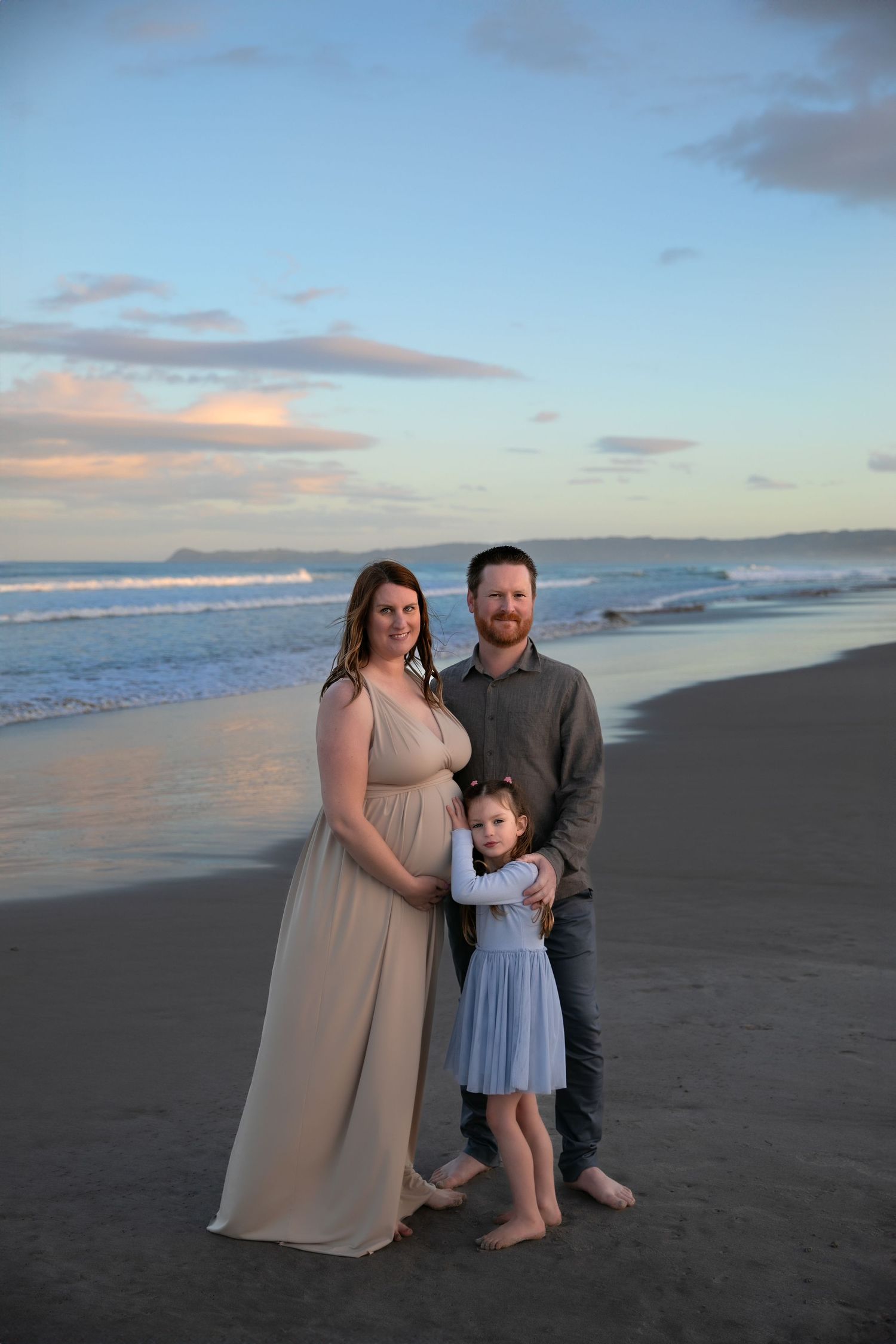 Family of three poses for sunset photos on a serene beach with waves and cloudy sky in the background.