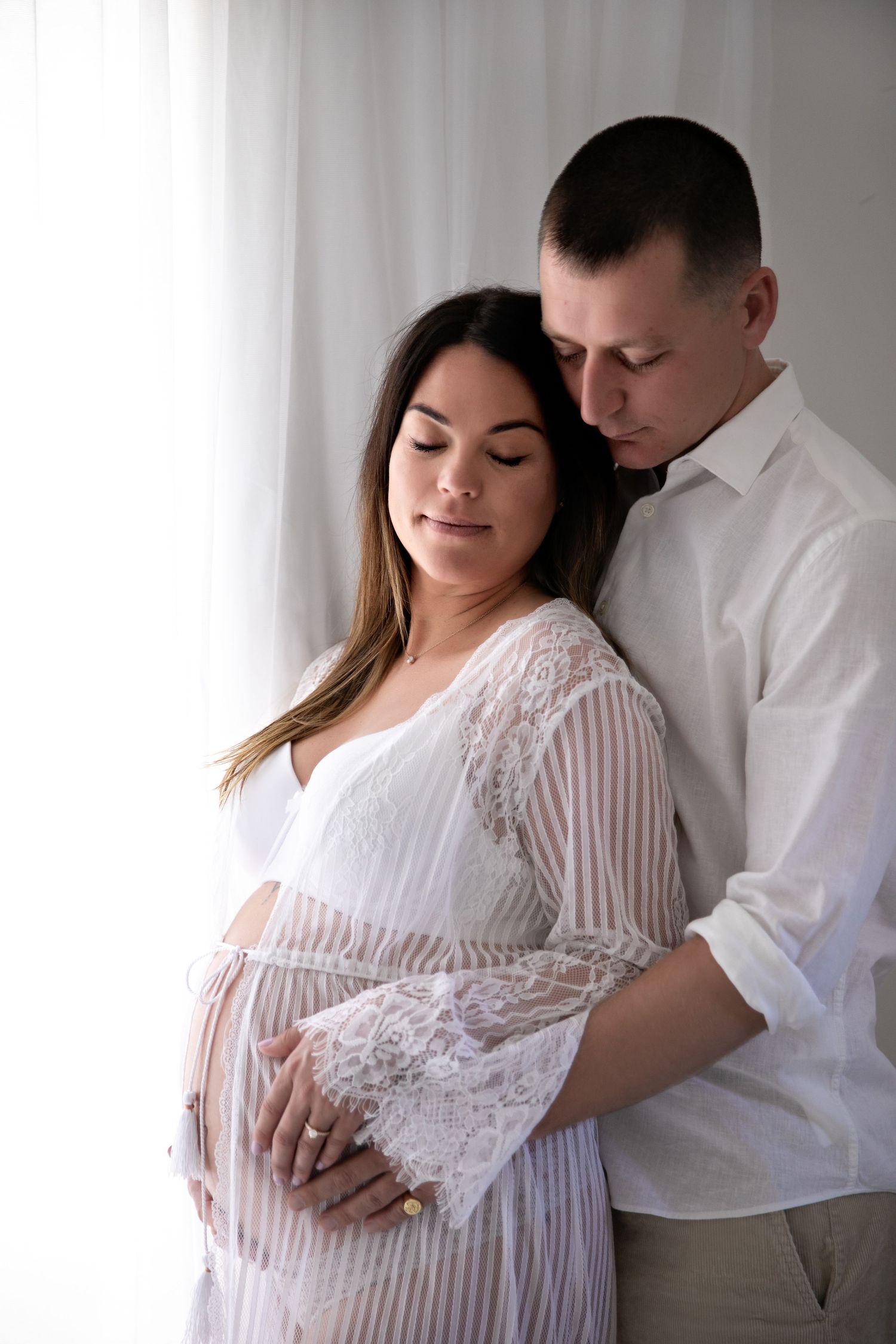 A couple in white embraces tenderly during a maternity photoshoot with soft lighting and neutral tones.