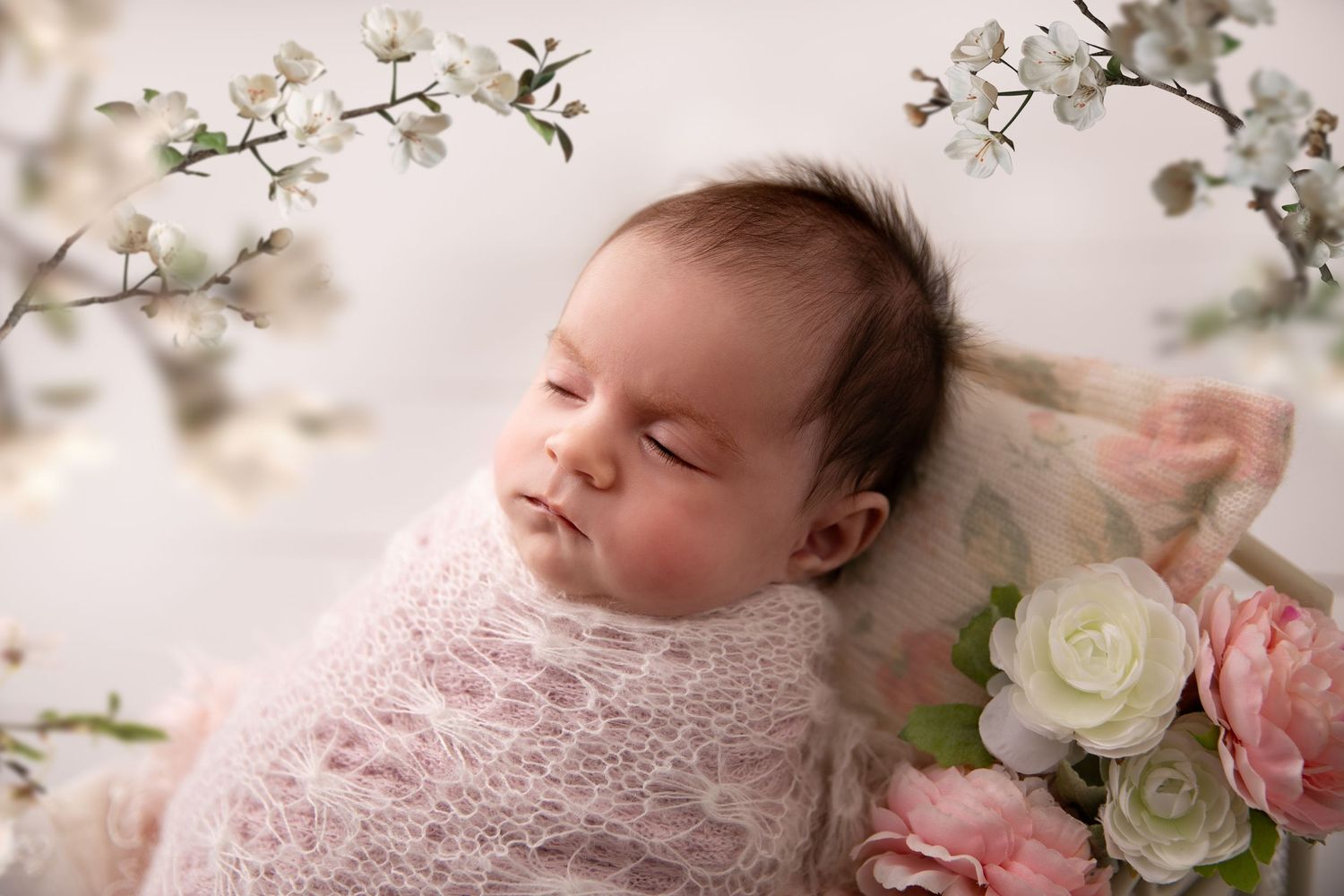 Newborn baby sleeping peacefully among delicate white cherry blossoms and soft pink floral arrangements.