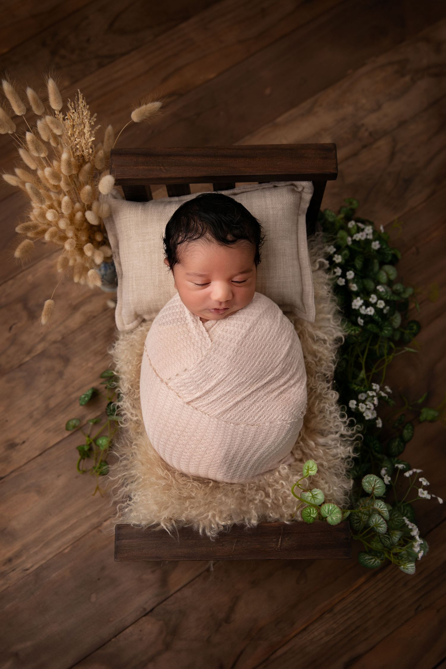 Newborn baby wrapped in soft pink blanket on wooden floor with delicate floral decorations and white accents.