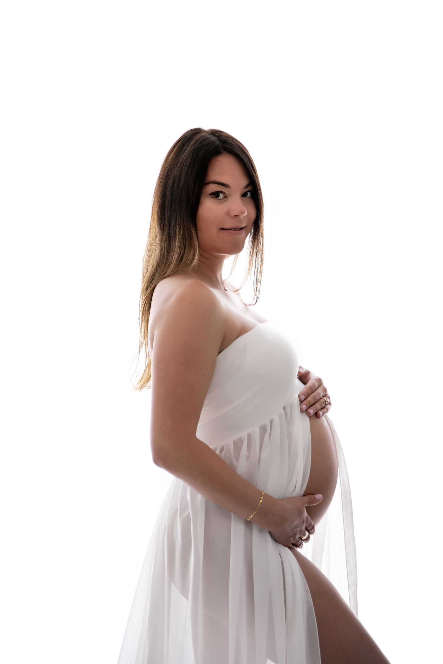 Elegant maternity portrait of an expectant mother in a flowing white dress against a bright background.